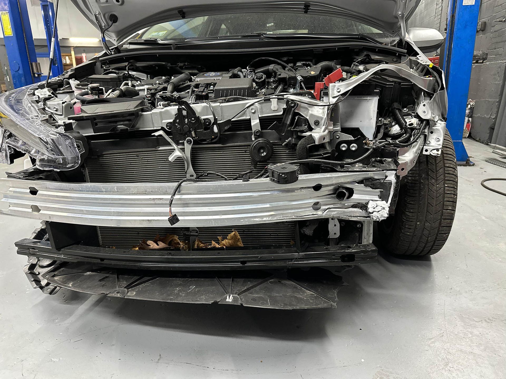 Damaged silver car in a garage with front end collision damage, hood open.