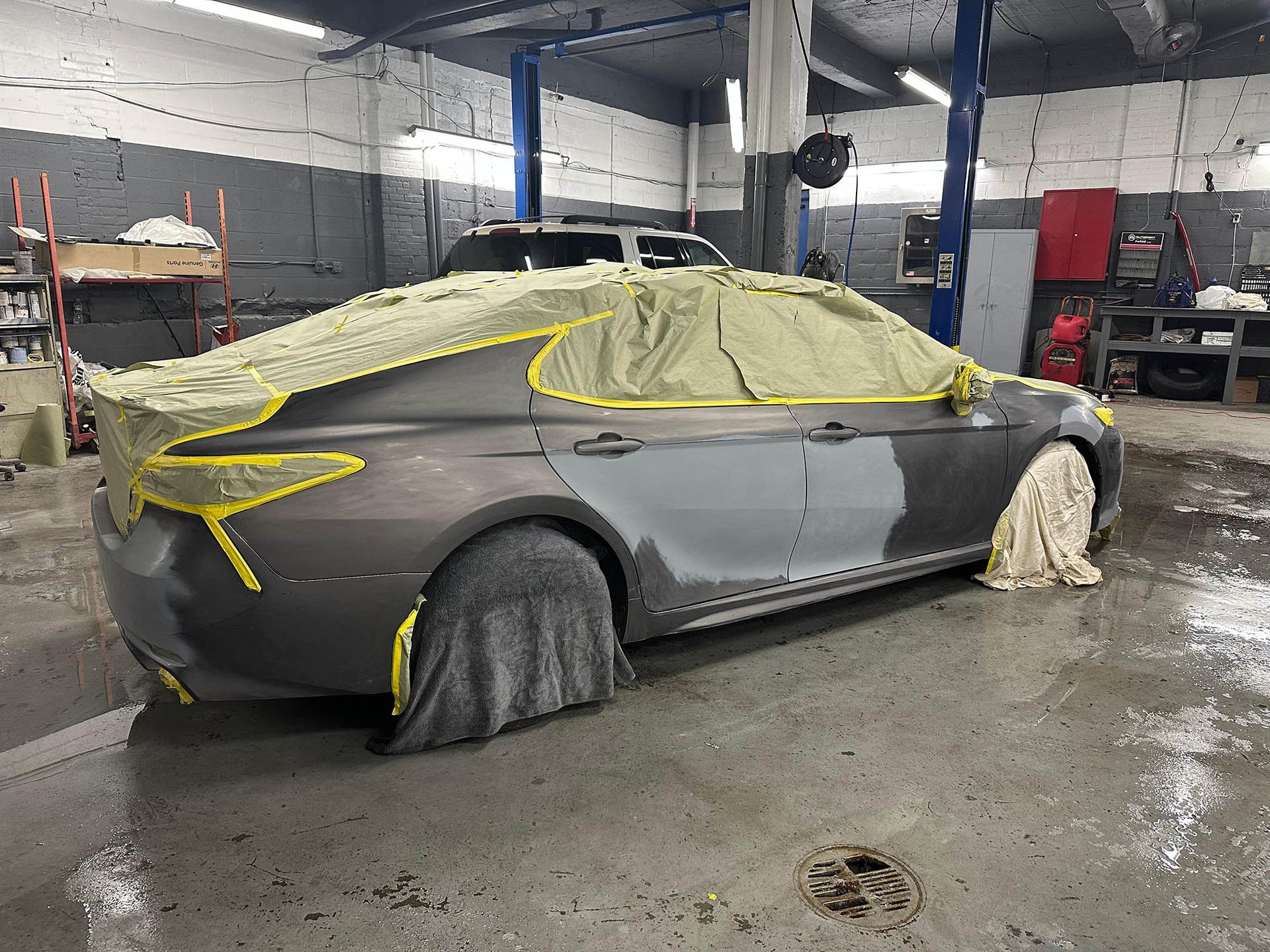 Car in auto body shop, partially masked for paint. Grey car has primed panels and is covered in yellow tape.