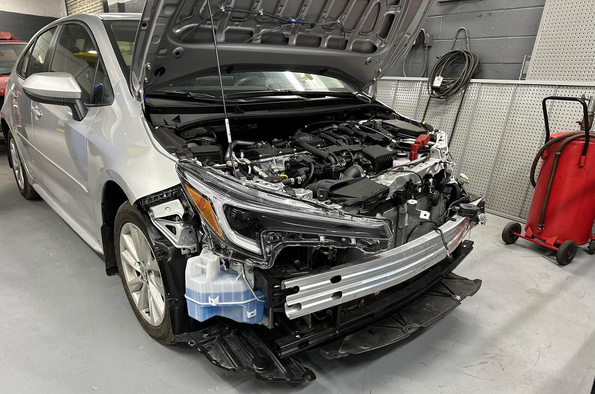 Silver car with front-end damage; hood open in a repair shop. Visible parts are crushed and broken.