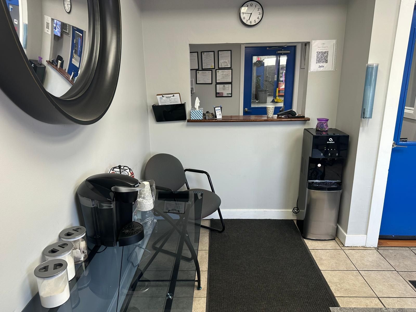 Waiting area in an auto shop: coffee station, desk, water cooler, clock, window to workspace.