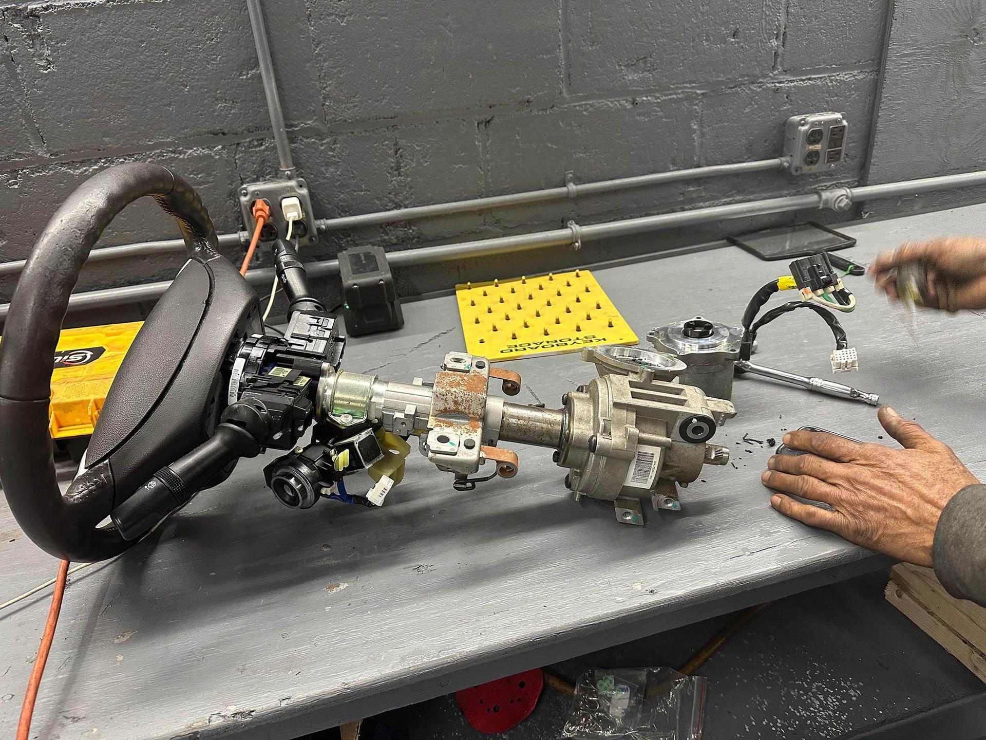 Steering column assembly on a workbench, with a hand reaching towards it.