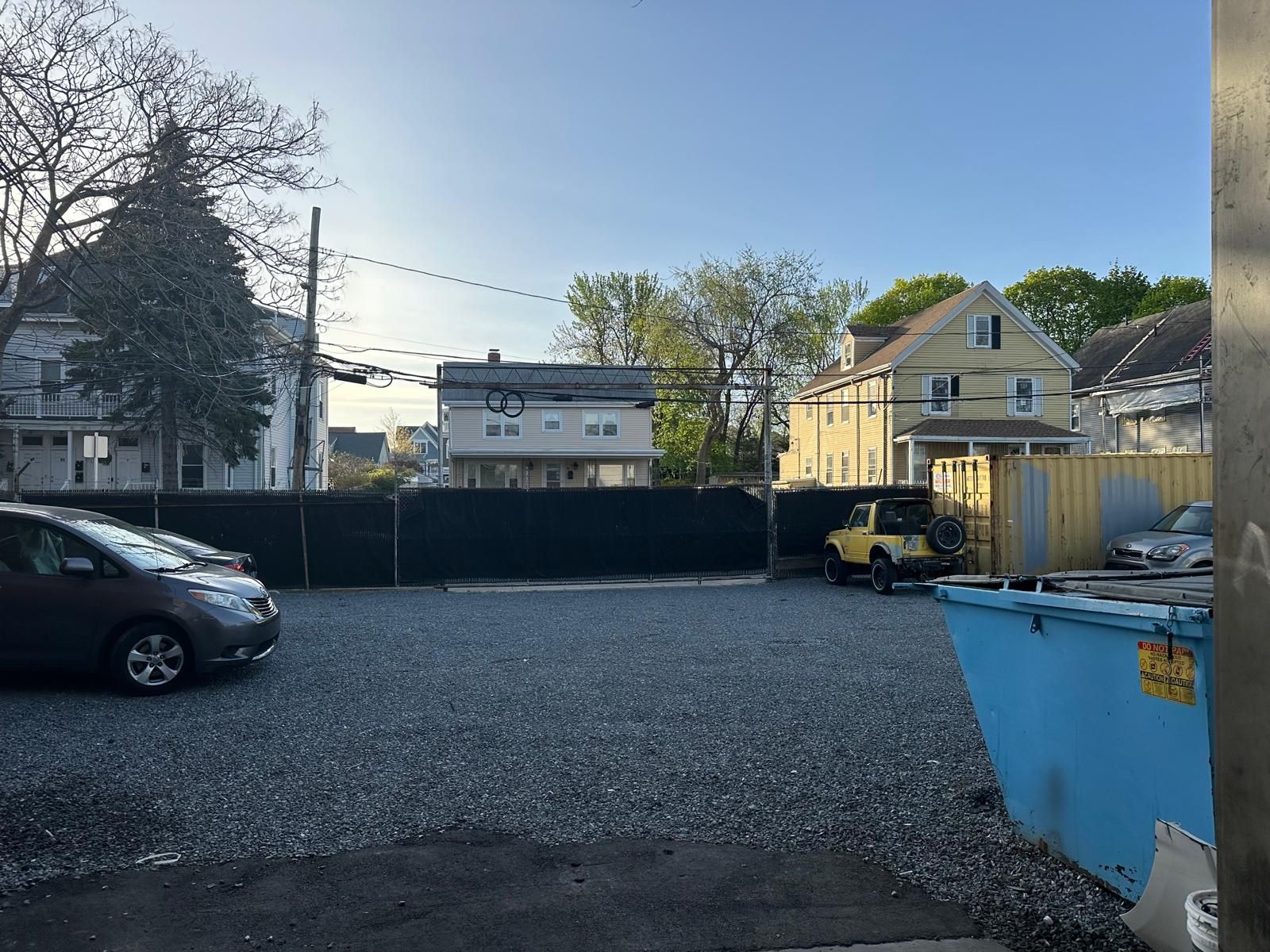 A gravel parking lot with parked cars, a yellow Jeep, and a large blue dumpster, set against a backdrop of houses.