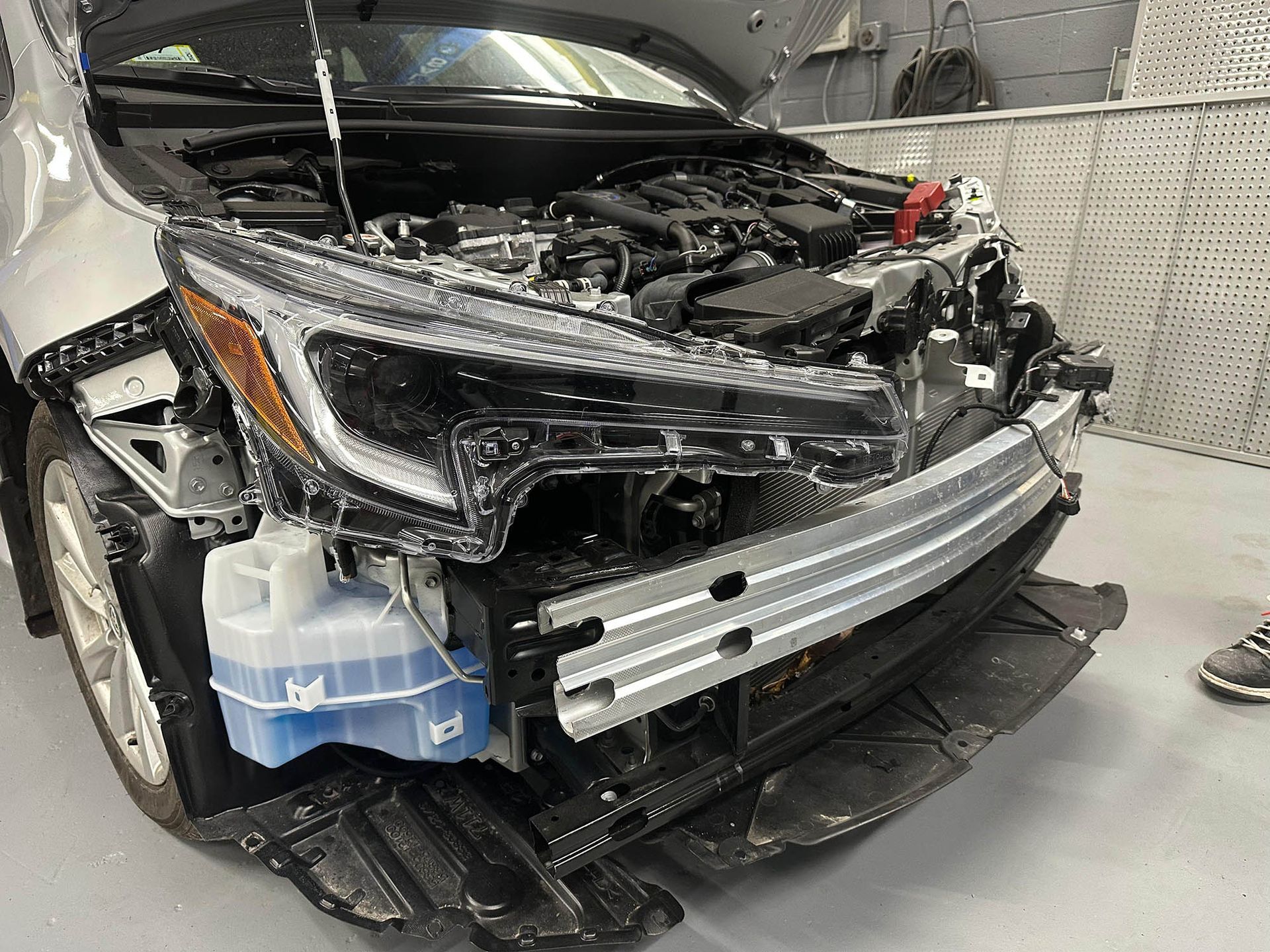 Silver car with front end damage, hood open in a repair shop.