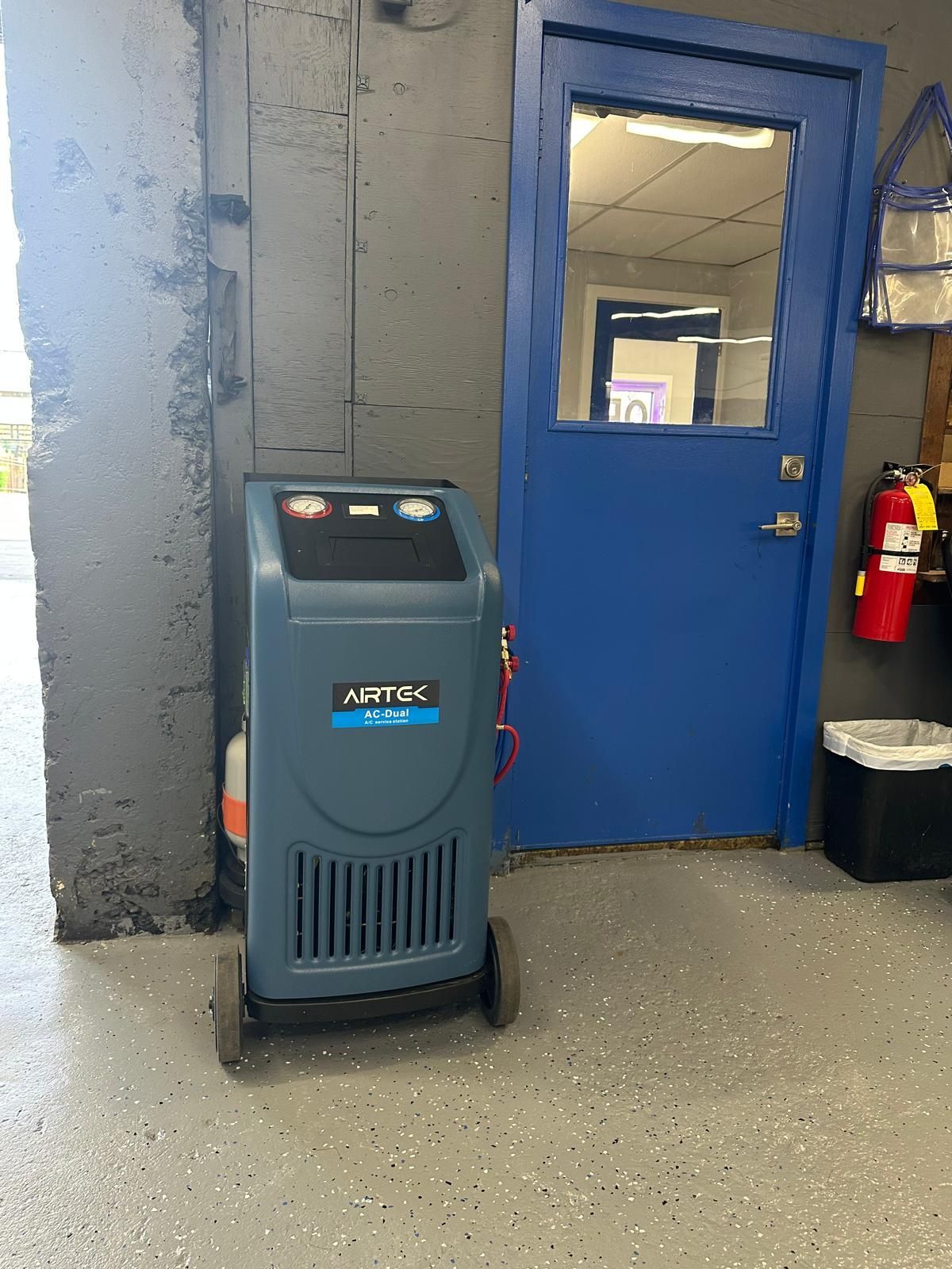Blue A/C service machine next to a blue door and gray wall. Fire extinguisher and trash can are nearby.