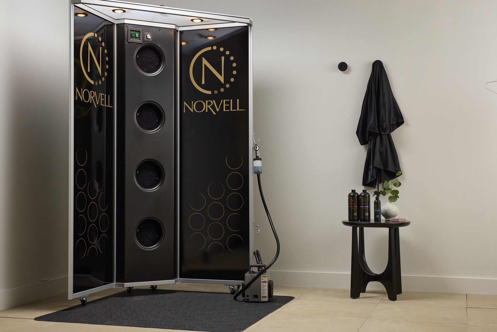 Sleek black Norvell tanning booth in a minimalist room beside a small table with folded towels and products