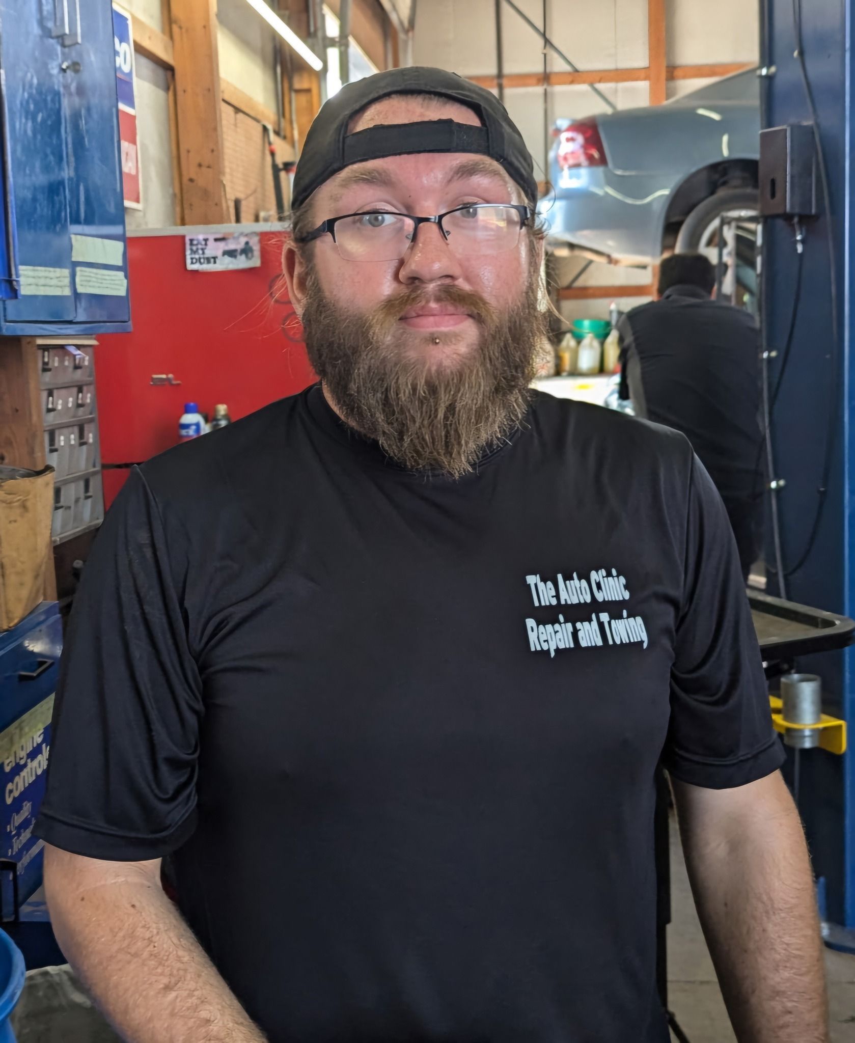 Man in glasses and cap, standing in a repair shop, wearing a black shirt with logo 