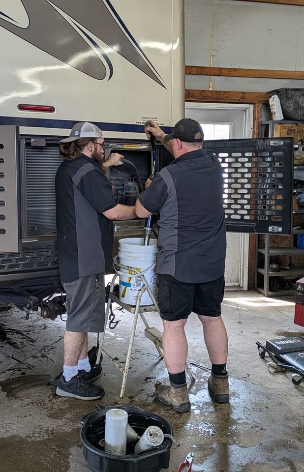 Two mechanics working on a recreational vehicle. One holds a part over a bucket, others in shorts and work shirts.