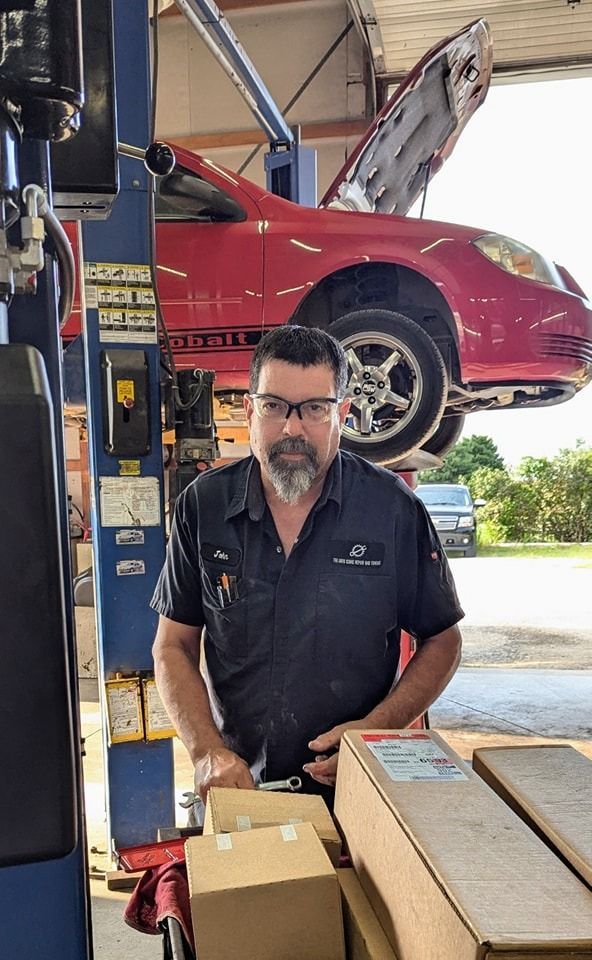 Mechanic in dark shirt stands in a garage, red car raised on a lift in the background, boxes in foreground.