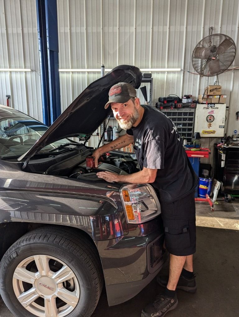 Mechanic working on a car with the hood up inside a garage. He’s wearing a hat, dark shirt, shorts.