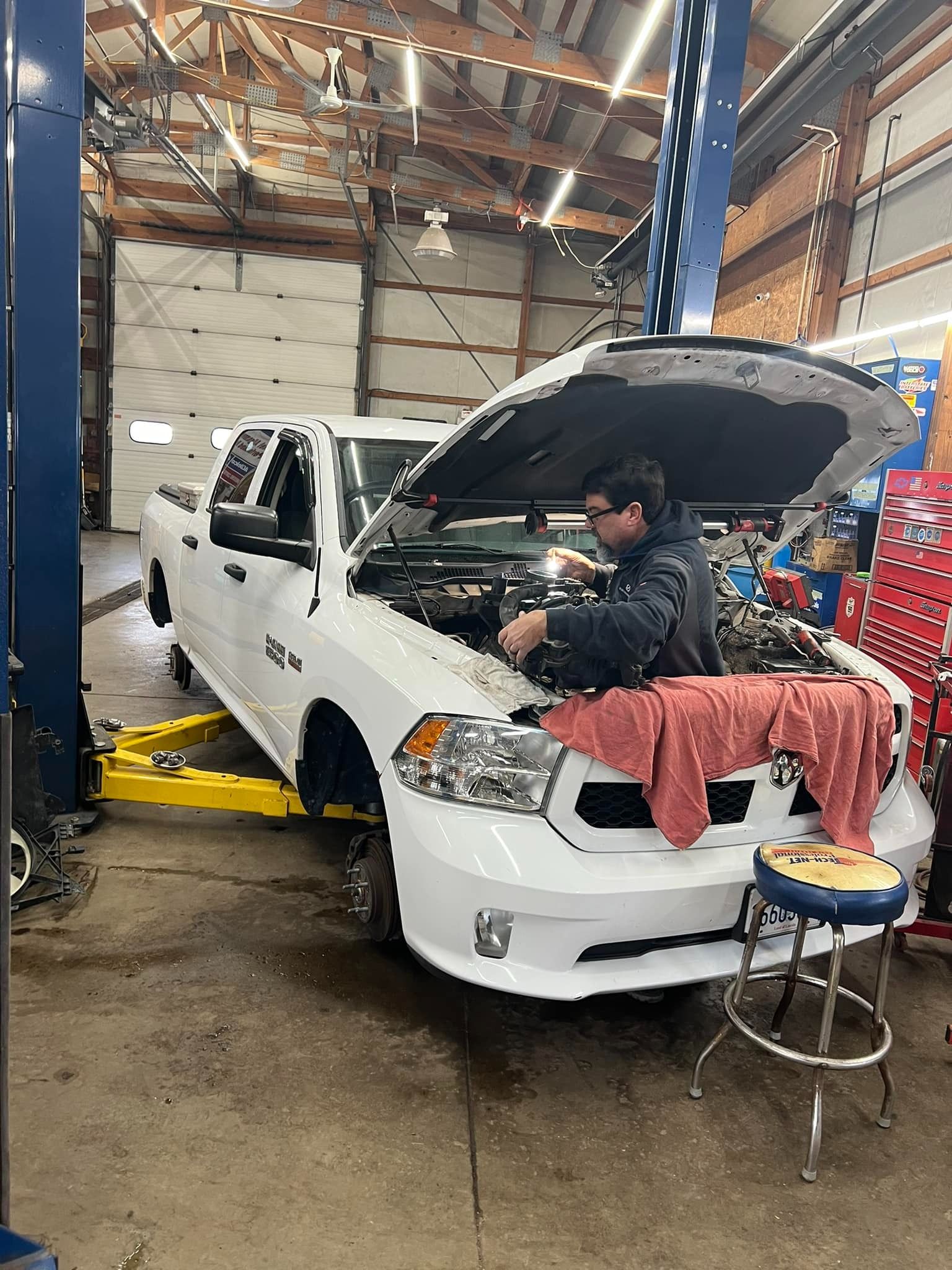 Mechanic working on a white pickup truck with its hood up in an auto repair shop.