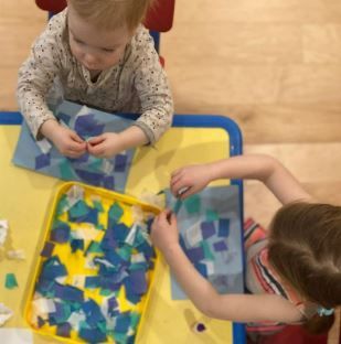 Two children are sitting at a table with a tray of blue and green papers