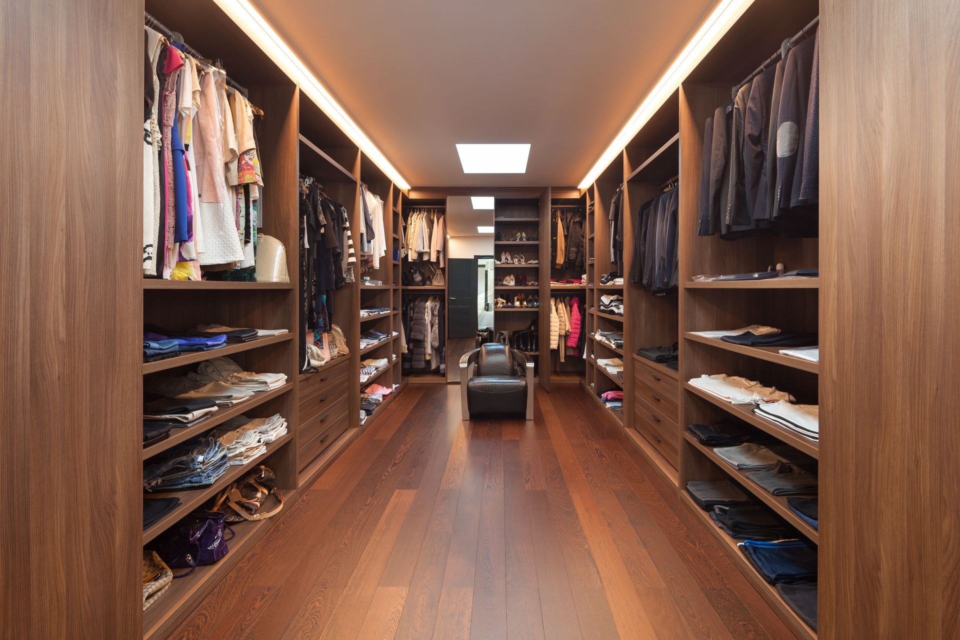 Walk-in closet with wood shelving, hanging clothes, folded items, and a central bench under warm lighting