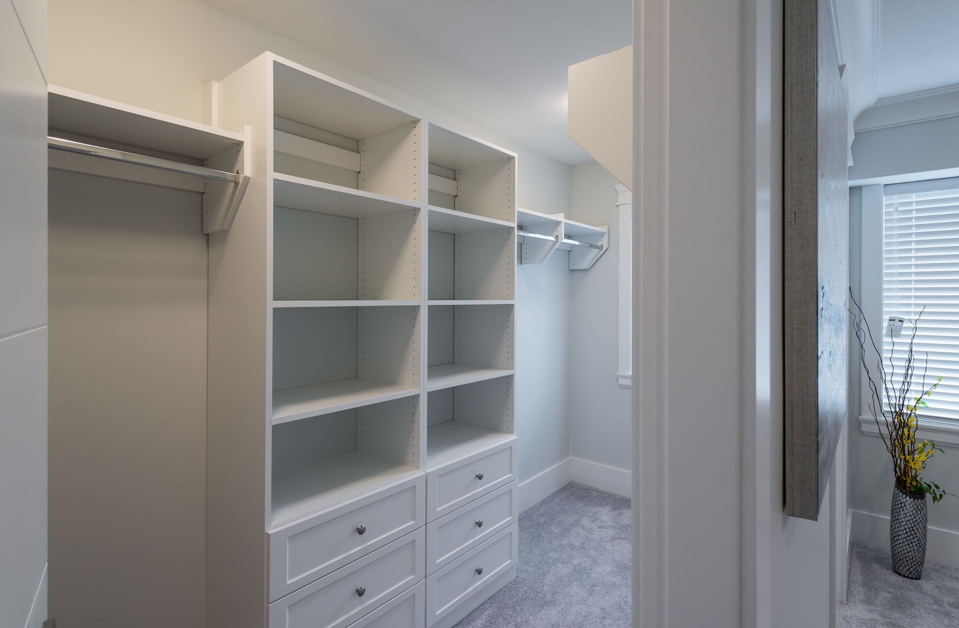 Bright walk-in closet with white shelves, drawers, and hanging space beside a bedroom window.
