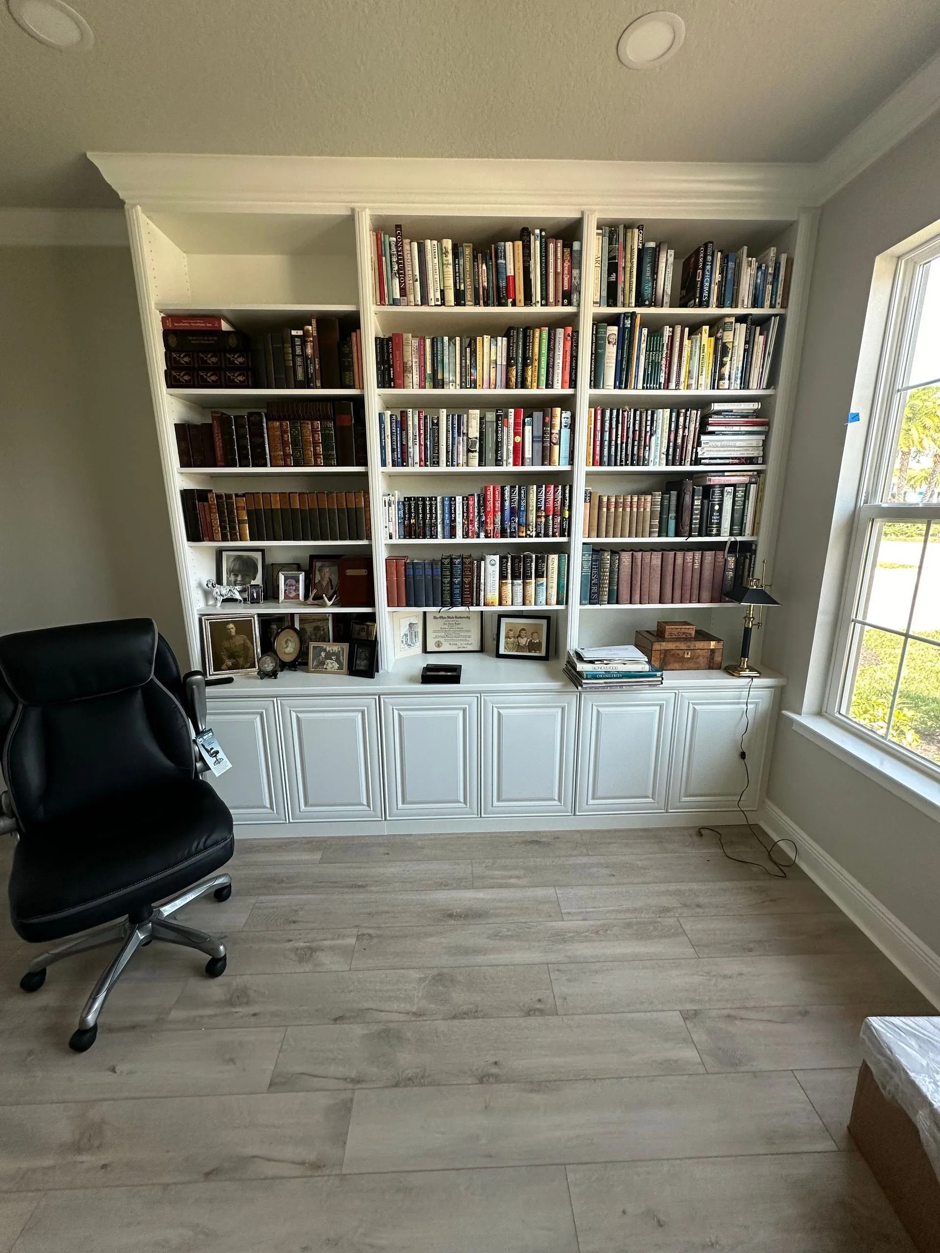 Home office with white built-in bookshelves, black chair, and large window with blinds