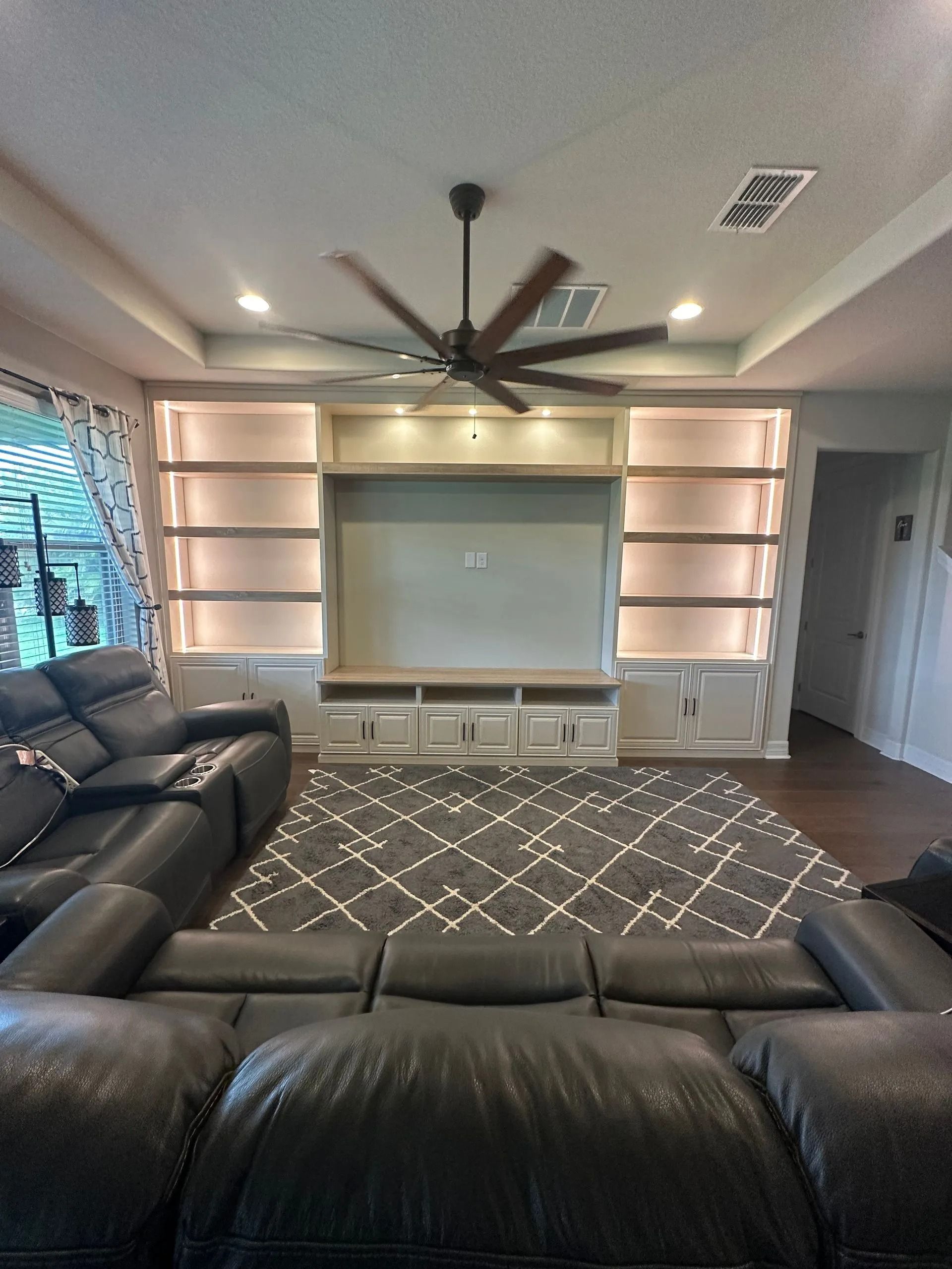 Living room with black leather sofas, ceiling fan, built-in shelves, and a large rug around a TV wall