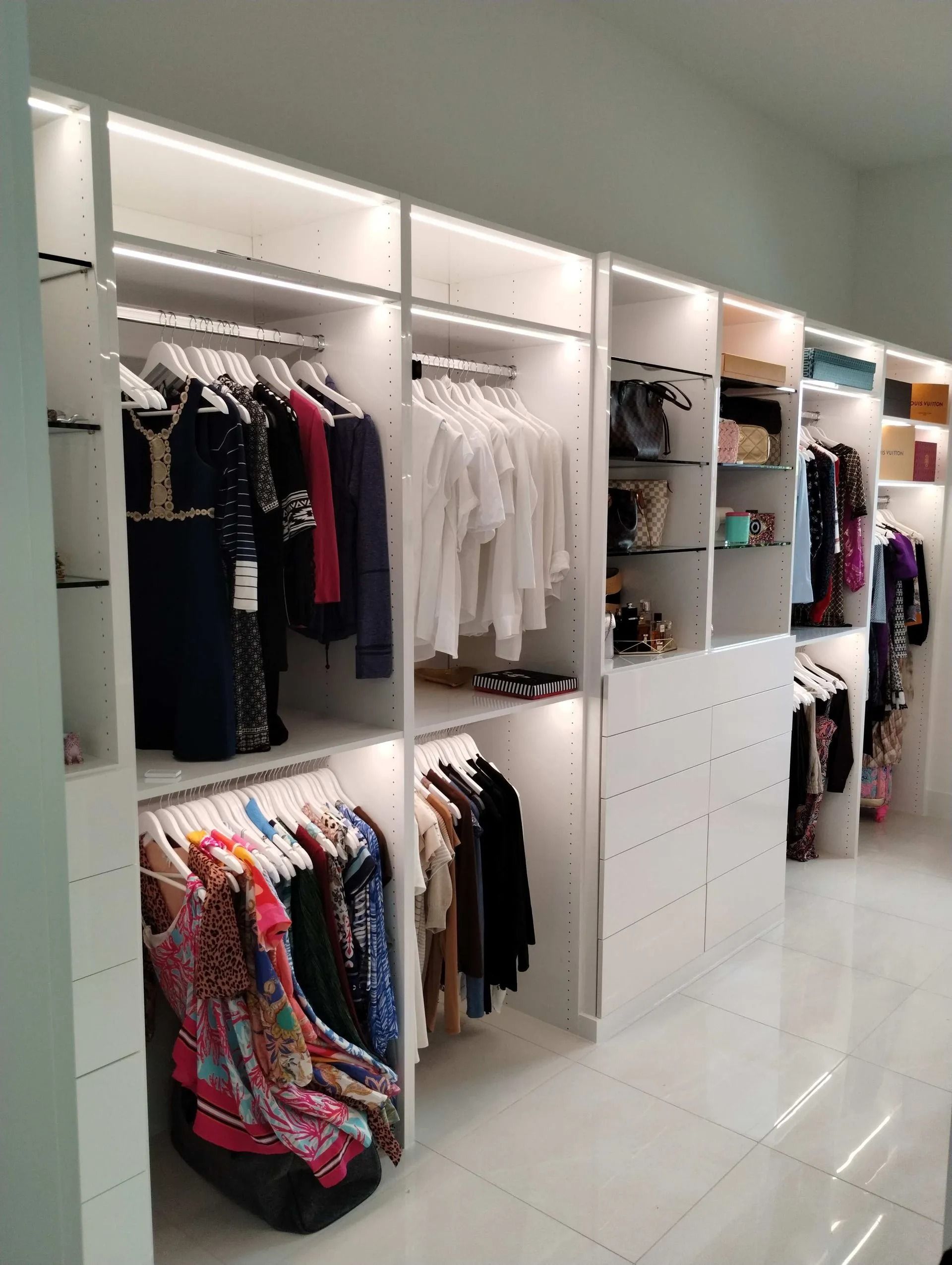 Bright walk-in closet with white shelves, hanging clothes, drawers, and a glossy tiled floor