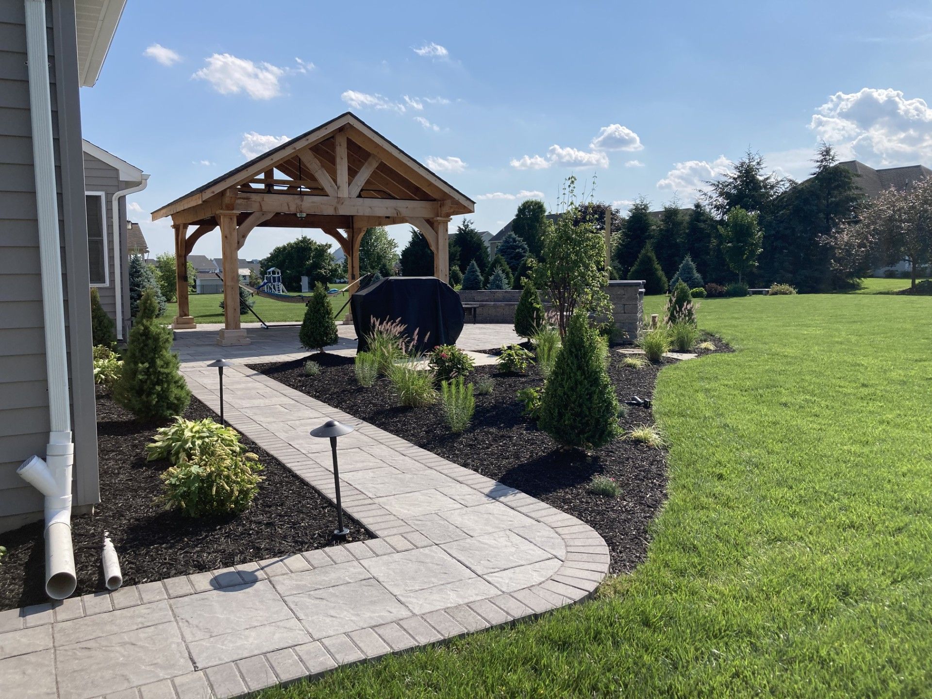 Backyard garden path leading to a wooden gazebo, with shrubs and a green lawn under a sunny sky