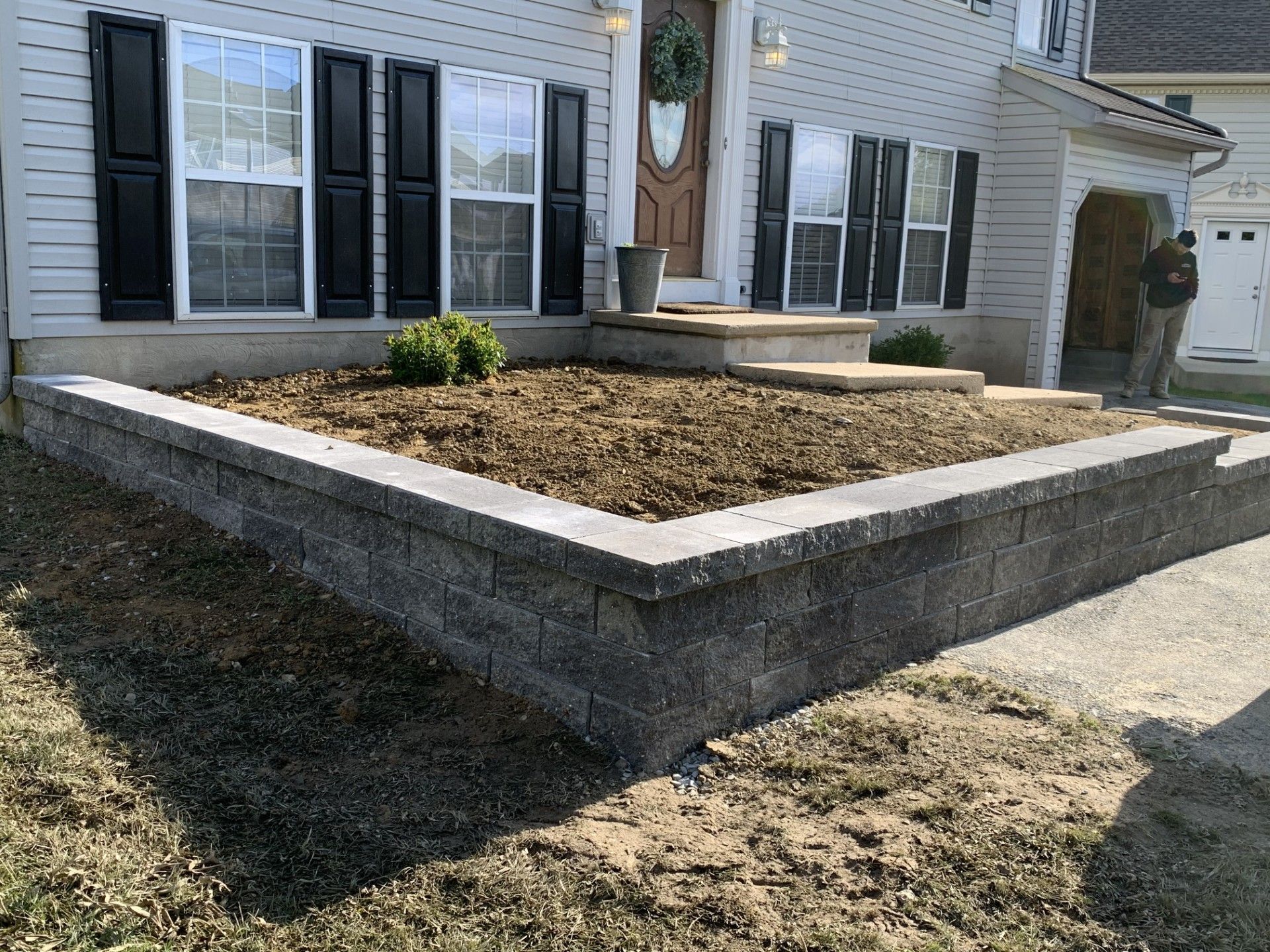 Raised front yard planter bed beside a white house with black shutters and a concrete retaining wall