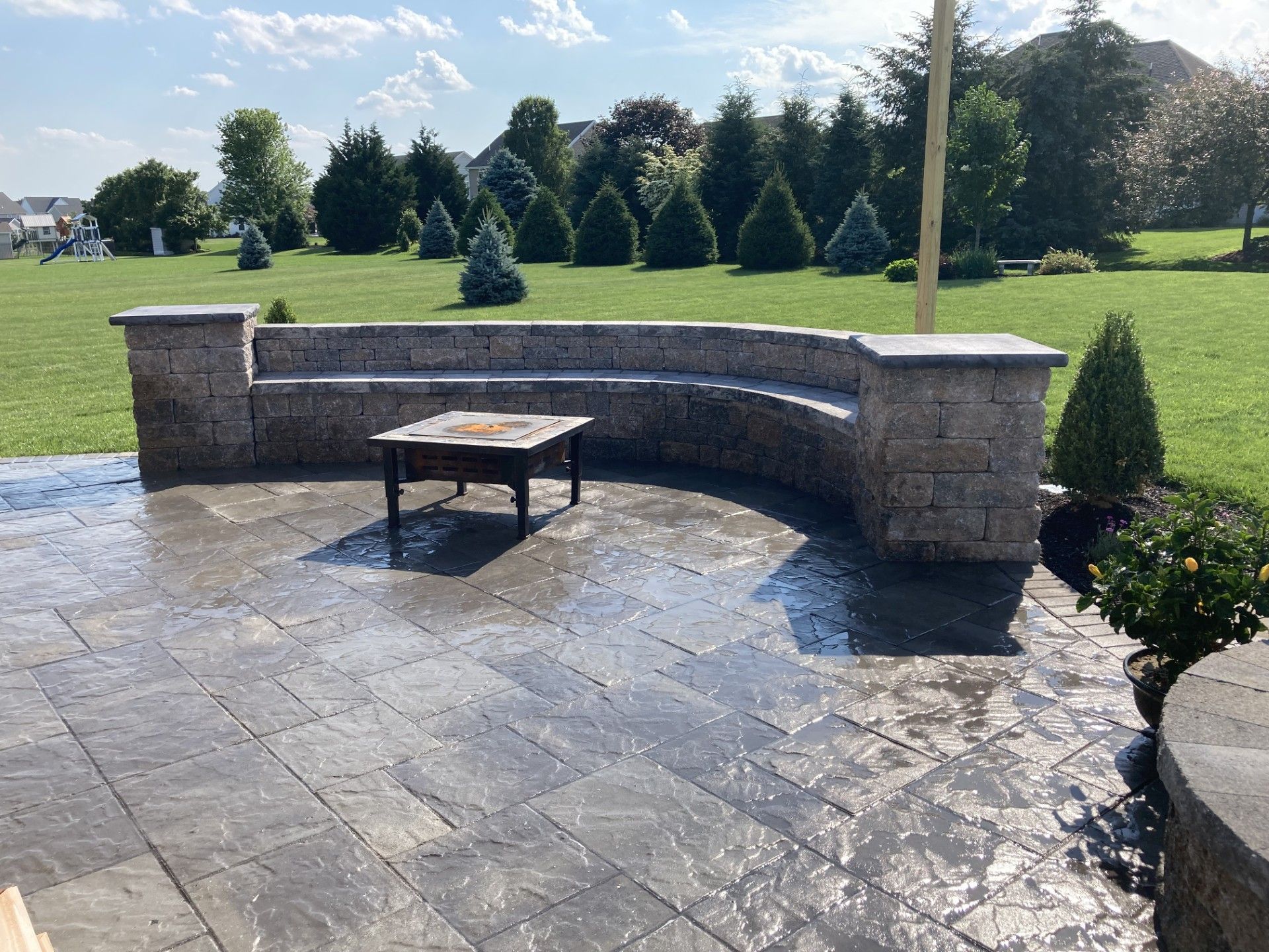 Stone patio with curved bench wall, small table, and sunlit grassy park in the background