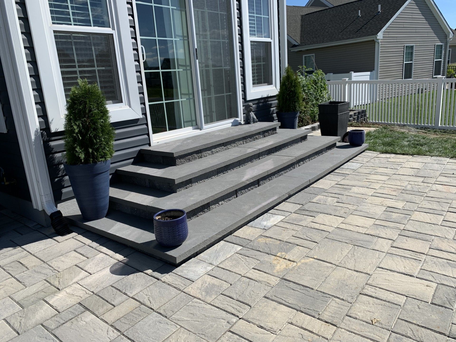 Front steps of a house with potted plants and a large paved patio in sunlight