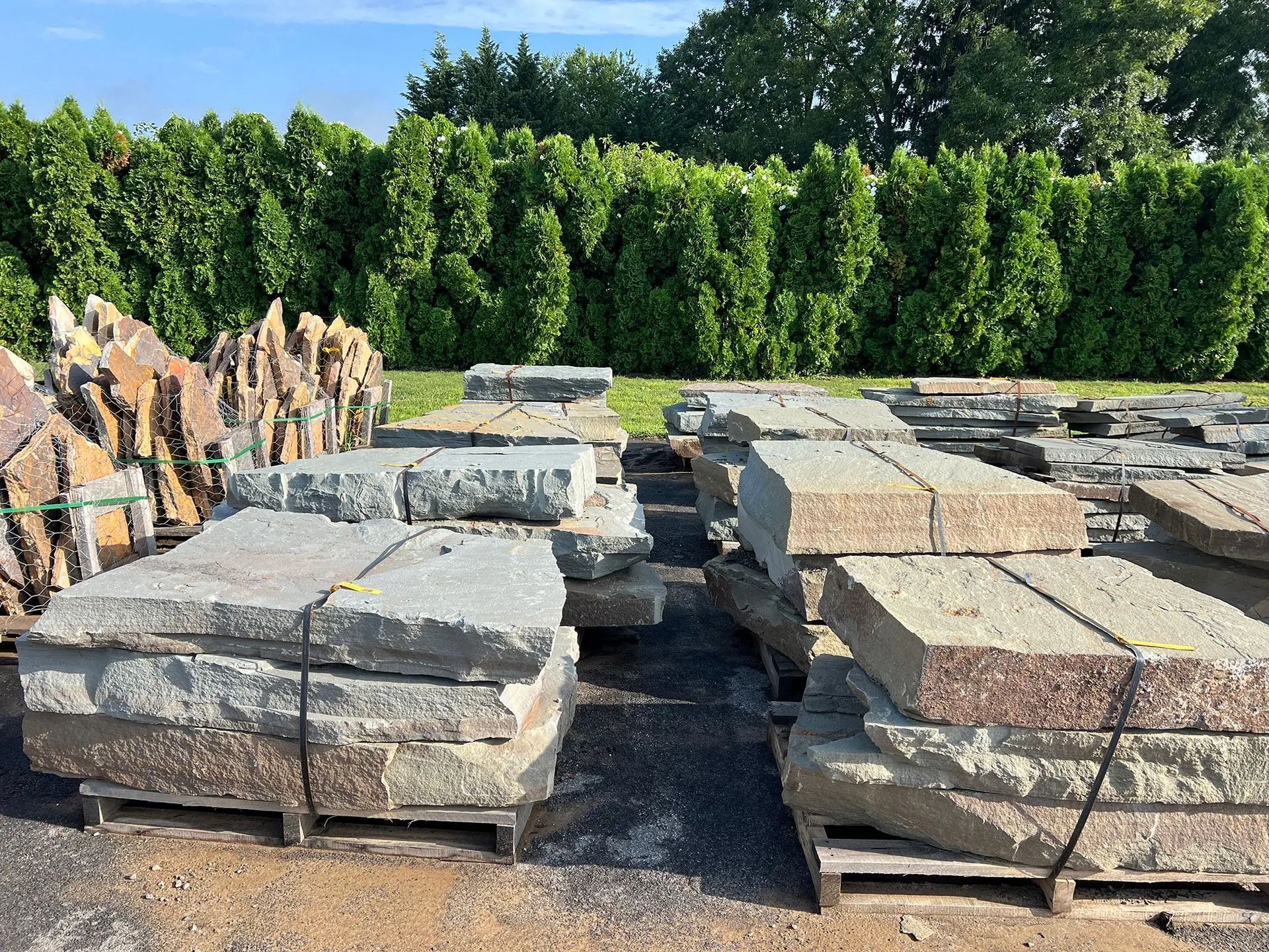 Stacks of stone slabs on pallets in a gravel yard with tall green hedges in the background