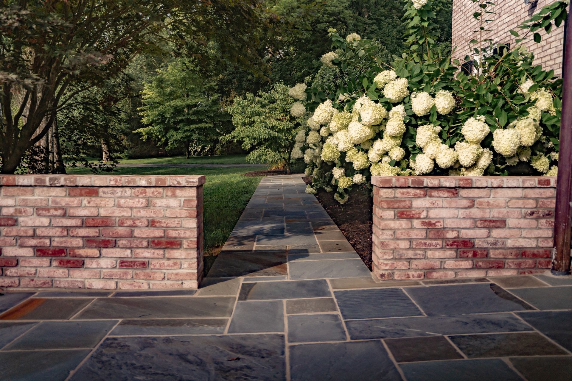 Brick garden entrance with stone walkway, brick walls, and white hydrangeas beside a house