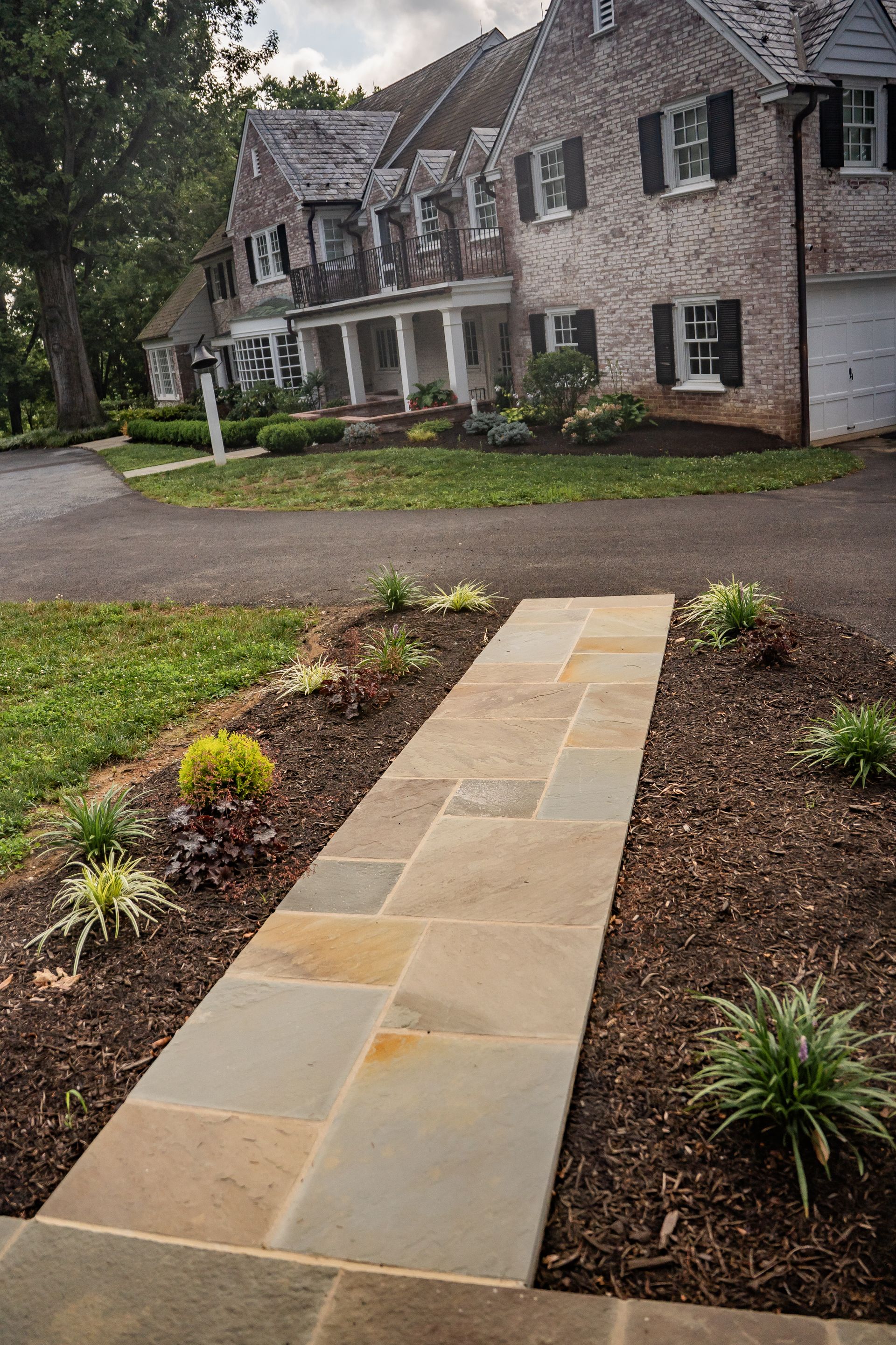 Stone walkway leads to a large brick house with a covered front porch and landscaped front yard.