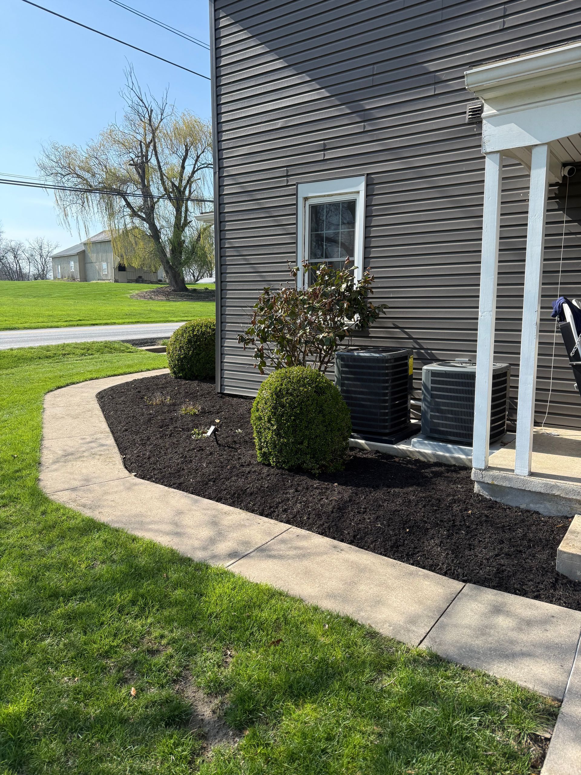 Corner of a house with a small landscaped bed, shrubs, and a curved stone border beside a sidewalk