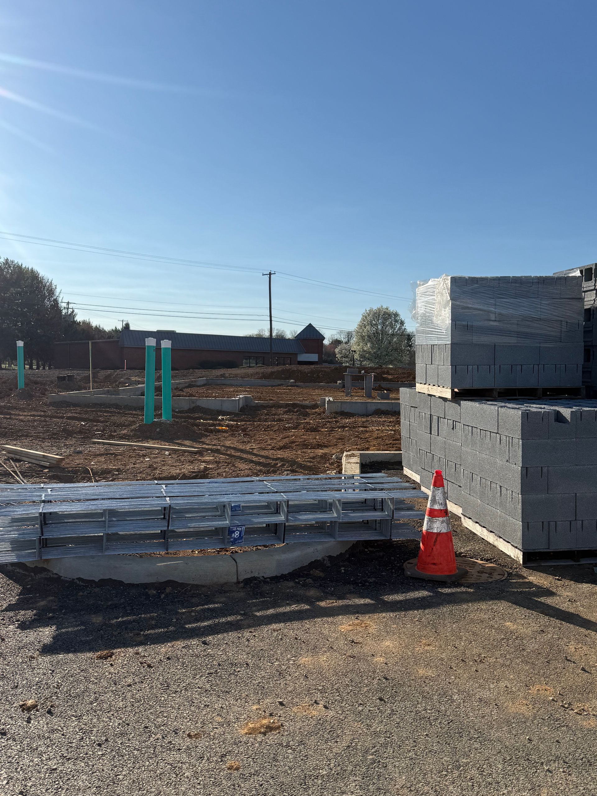 Construction site with stacked cinder blocks, orange cone, and metal materials on a gravel lot under a clear blue sky