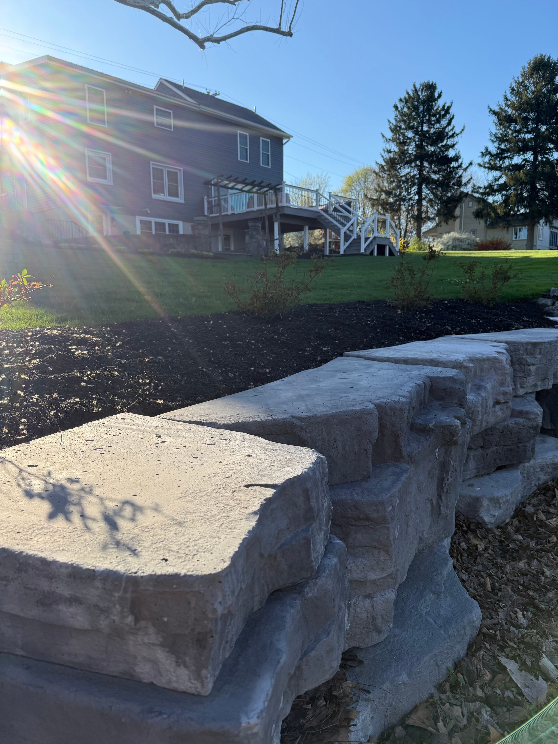 Stone retaining wall in foreground with a sunlit house and tree in the background.