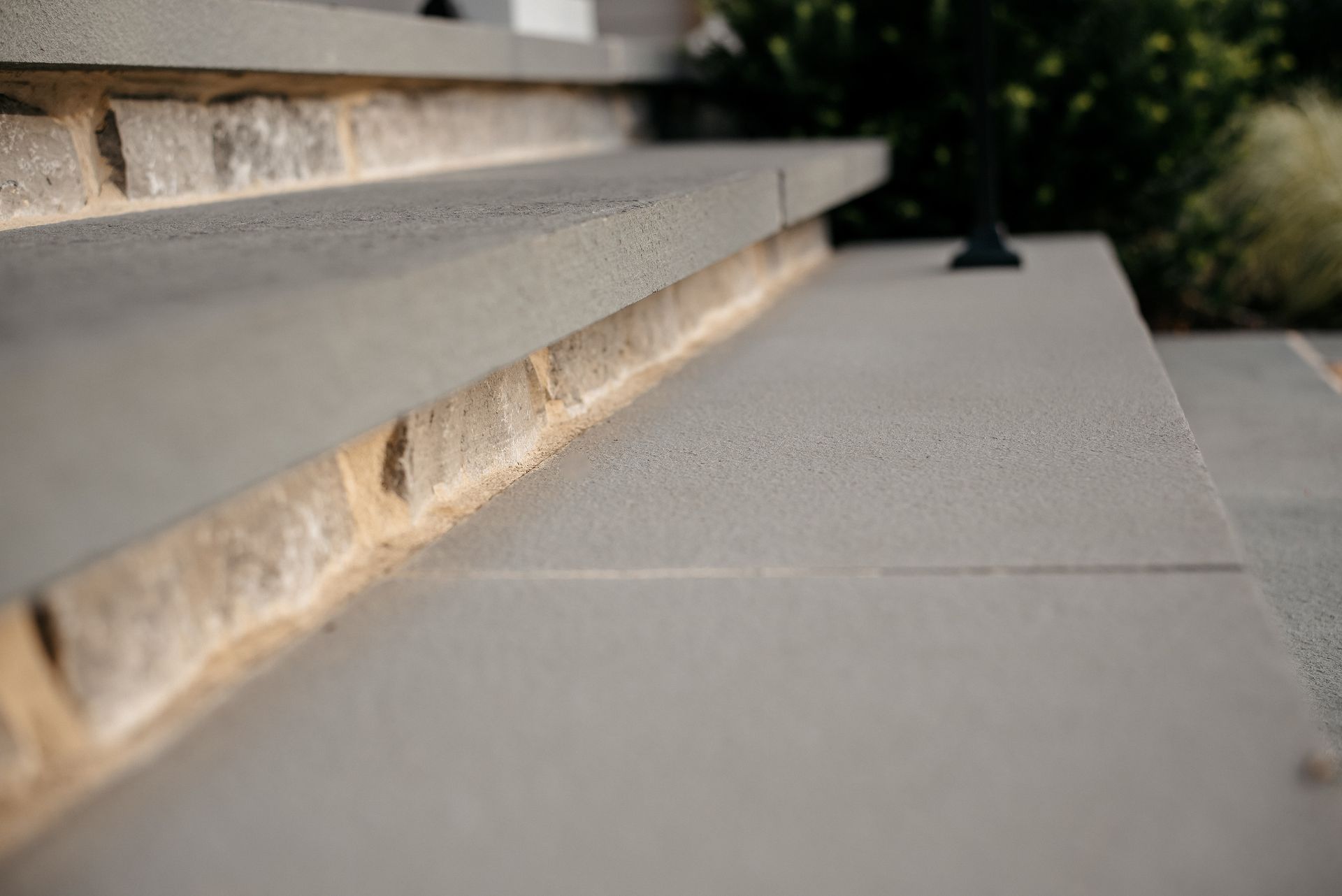 Close-up of a stone step and smooth concrete walkway beside a wall, with blurred greenery in the background