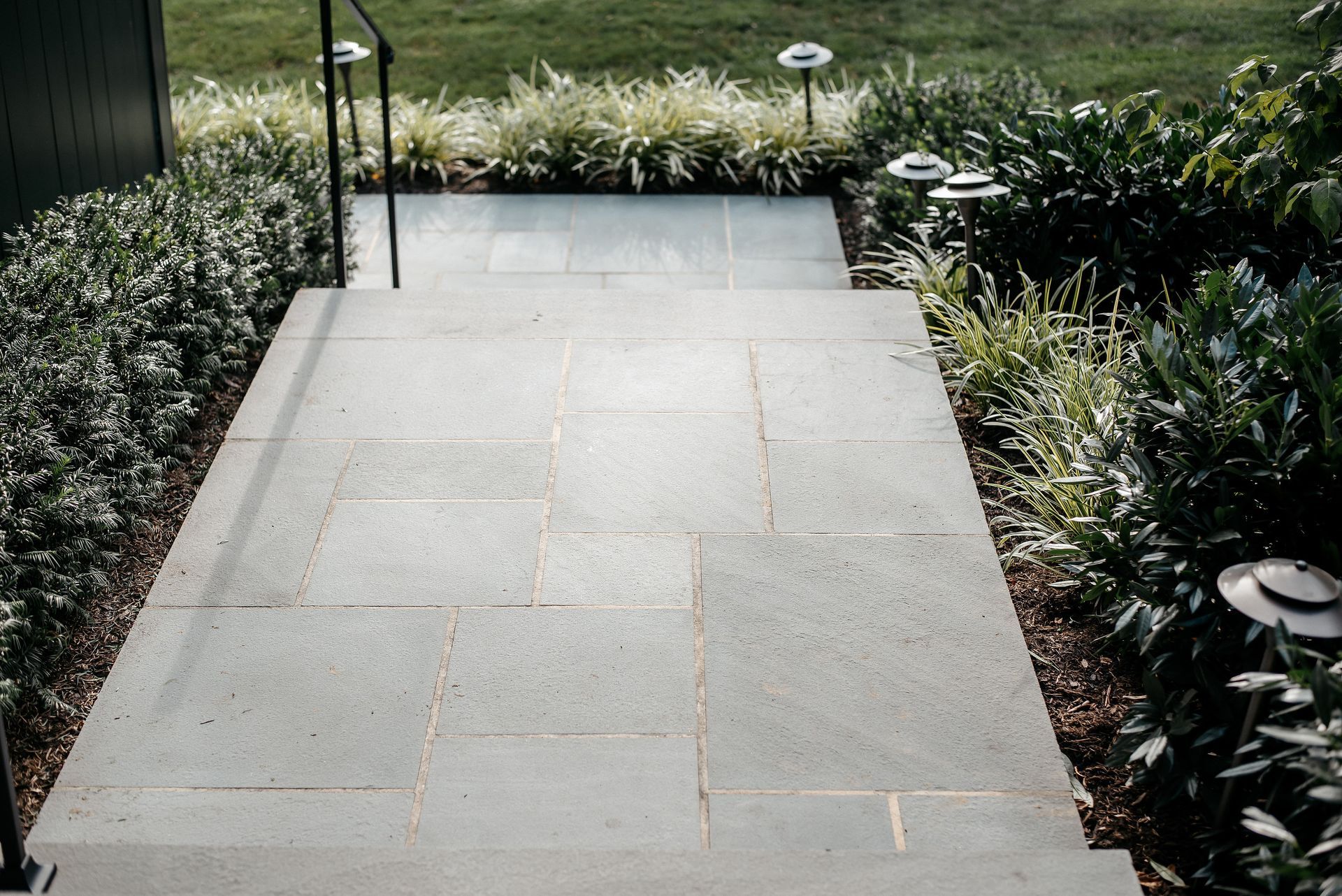 Stone garden pathway bordered by shrubs and small solar lights leading to a grassy lawn