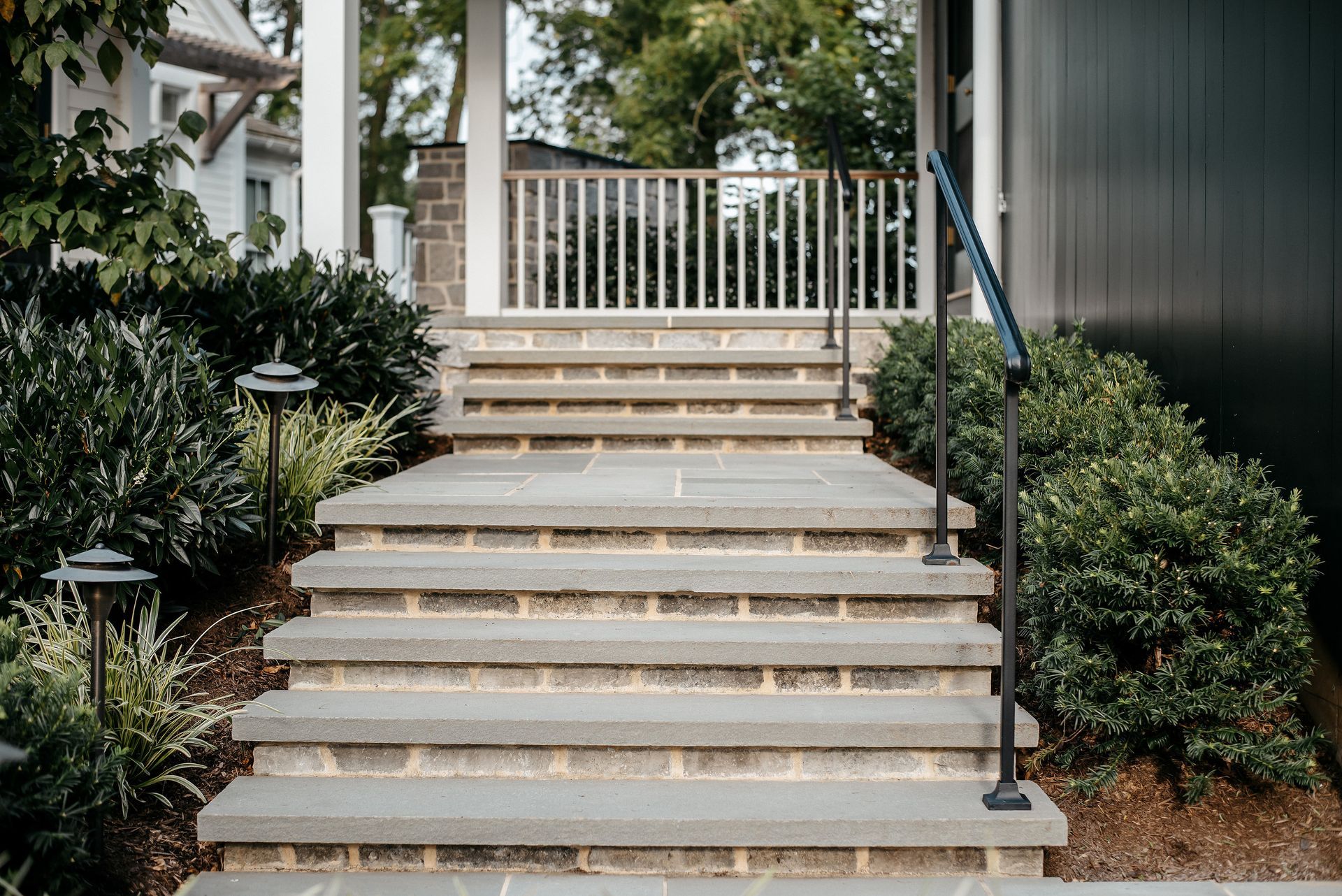 Front steps leading to a porch with white railing, surrounded by greenery and shrubs