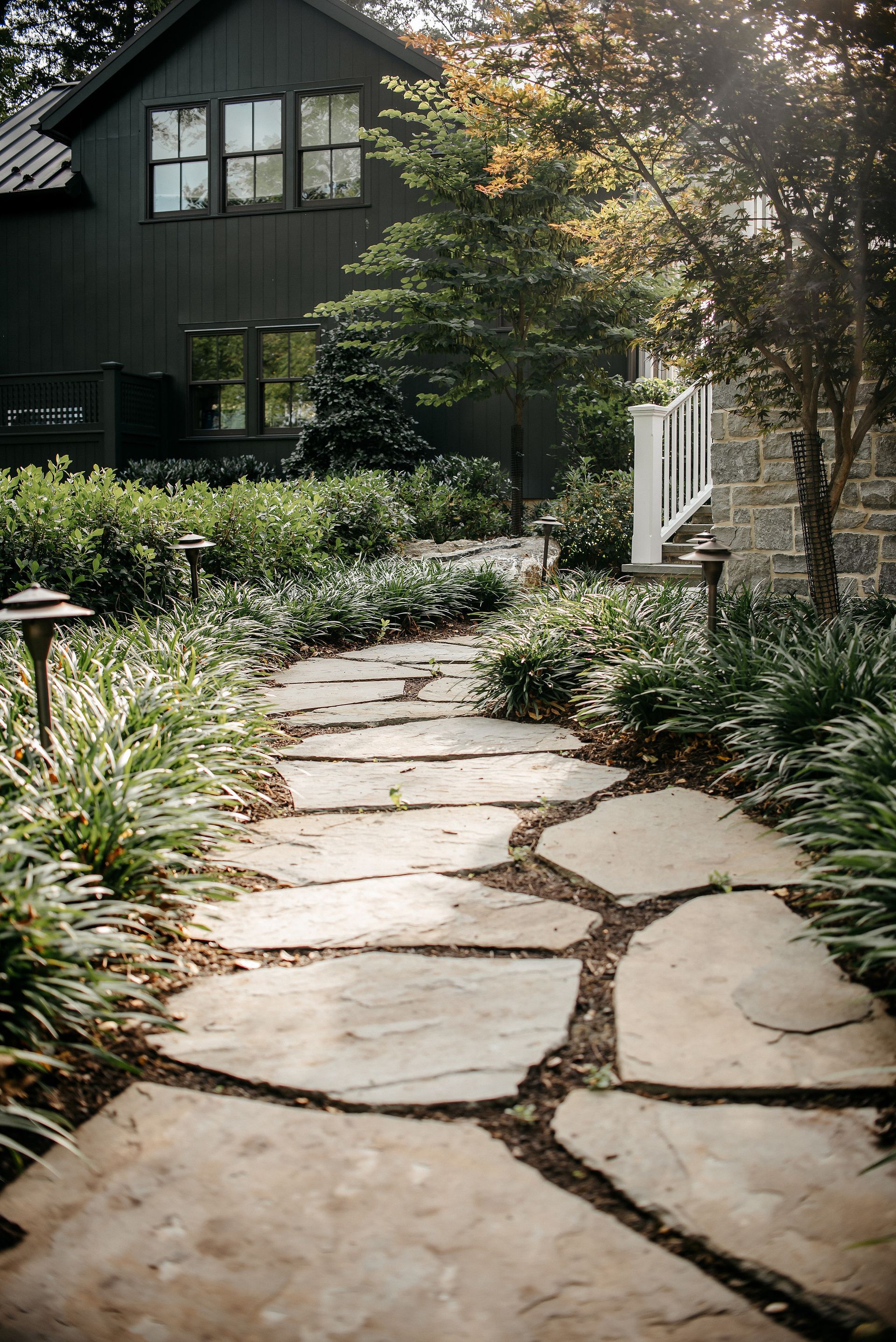 Stone garden path beside a house with lush greenery and sunlight filtering through trees
