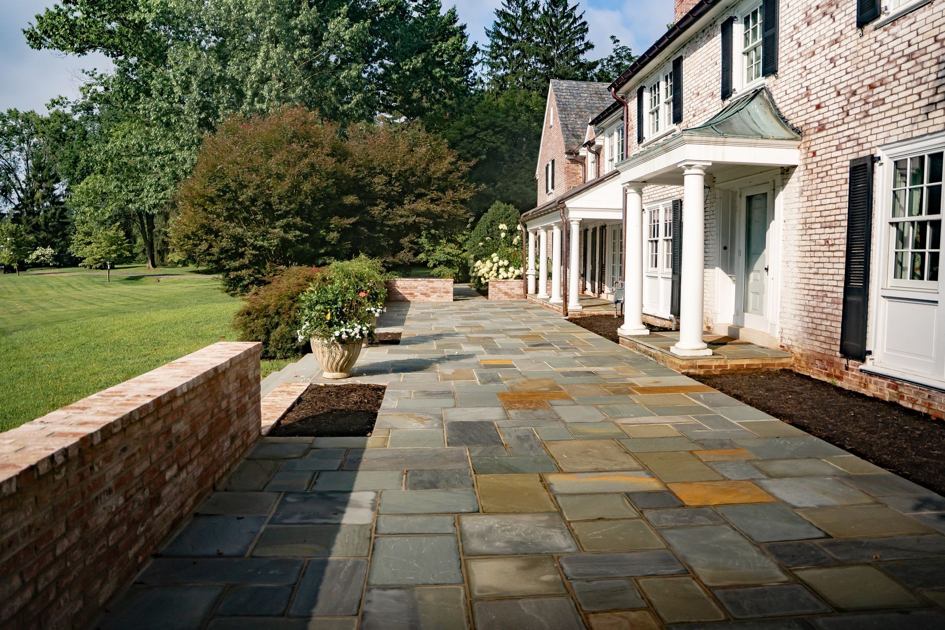 Stone patio beside a brick house with white columns, bordered by a low wall and green lawn.