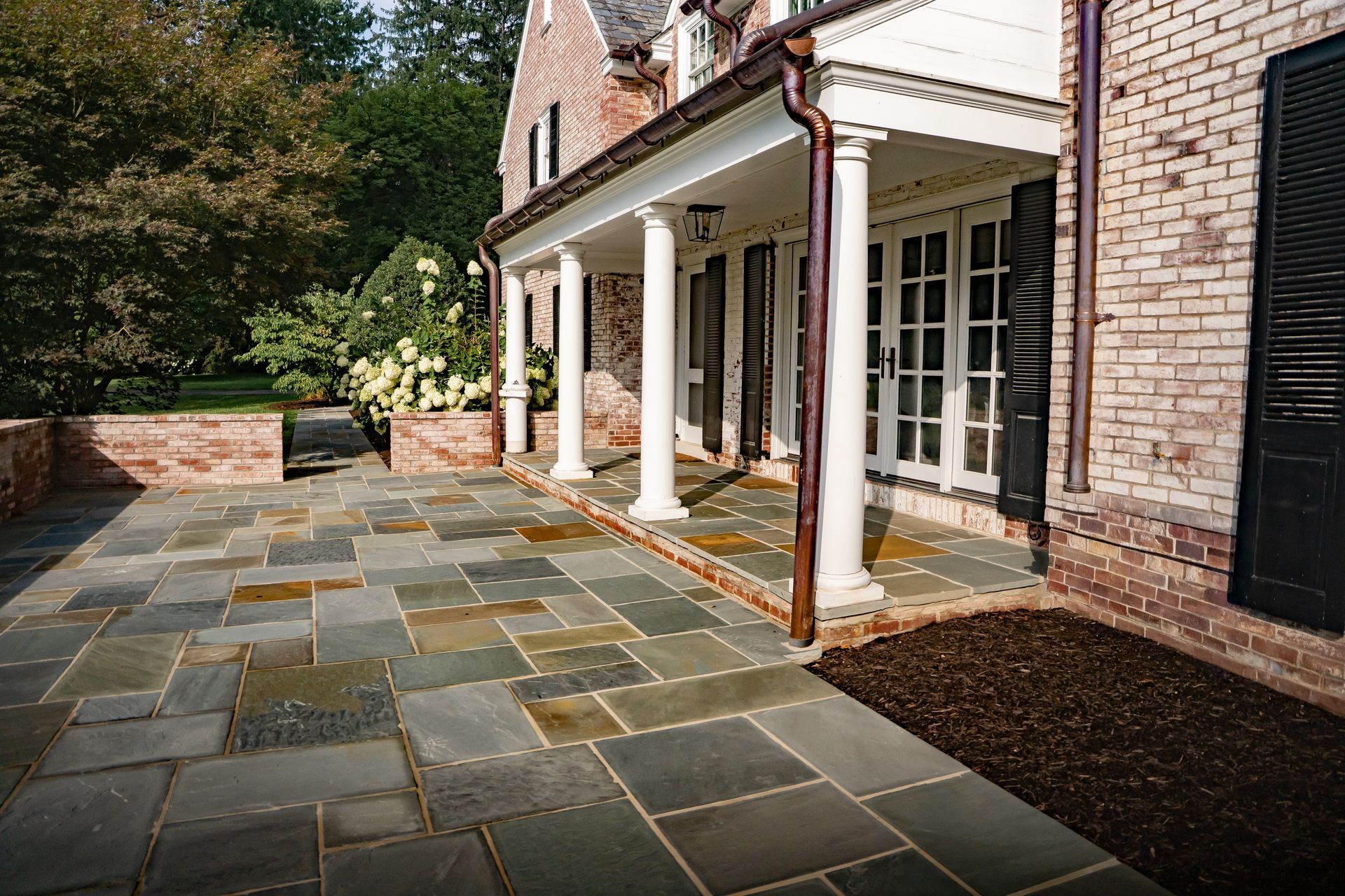 Stone patio beside a brick house with white columns and glass doors, bordered by trees and garden beds