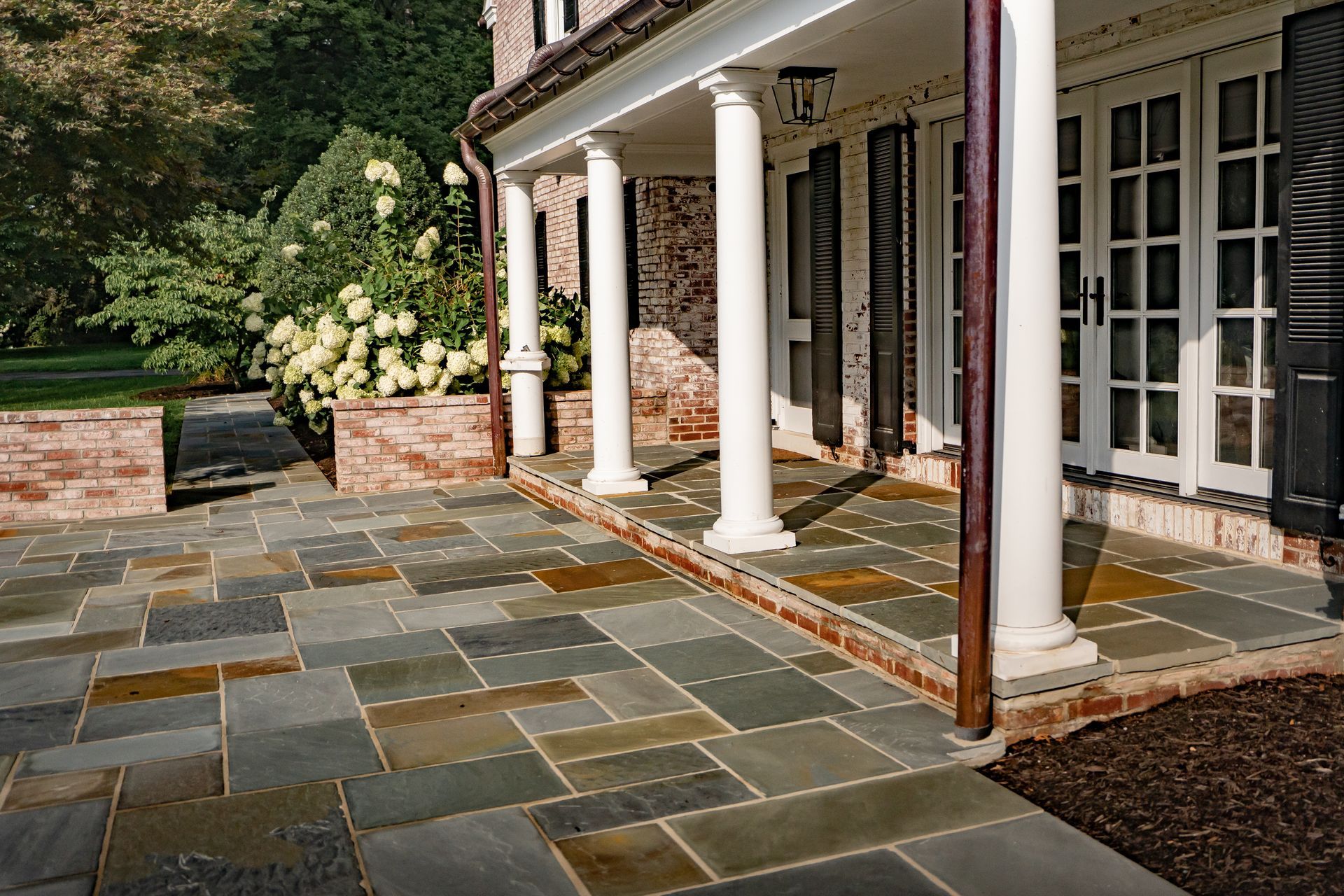 Stone patio beside a white-columned house with French doors and landscaped garden greenery