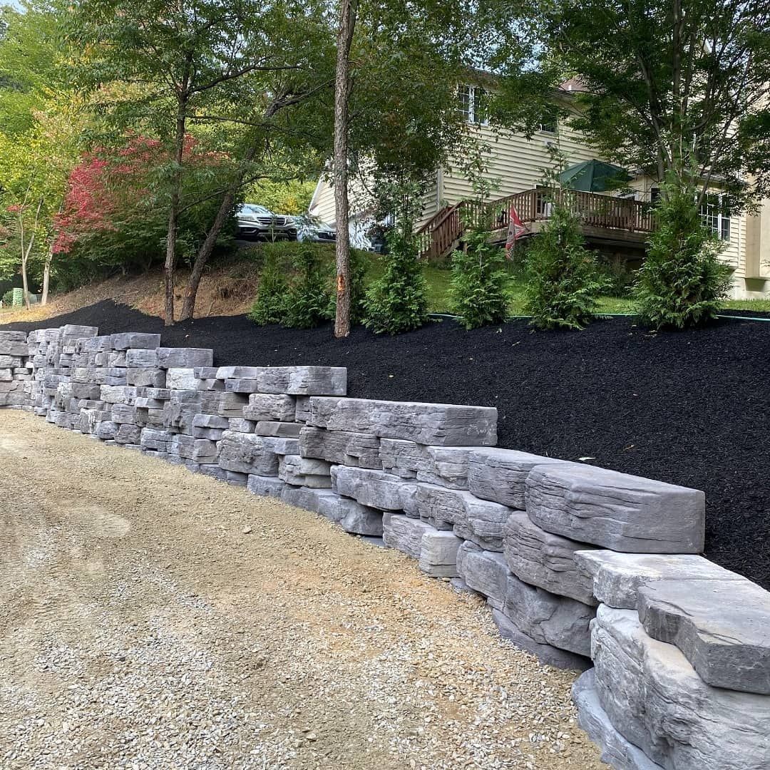 Stone retaining wall beside a sloped mulched yard with trees and a house in the background