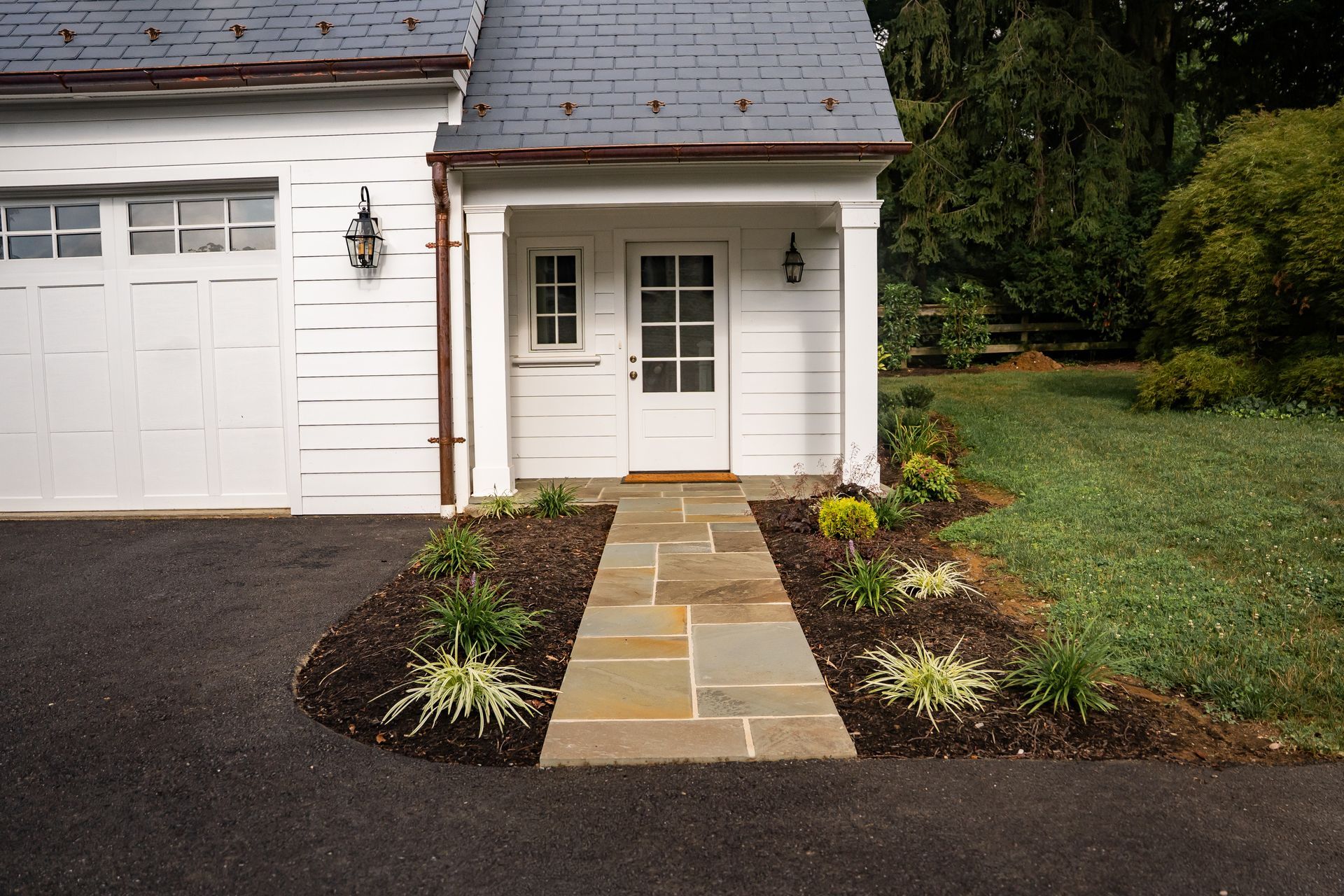 Front entrance of a white house with a stone walkway, garage door, and landscaped garden beds
