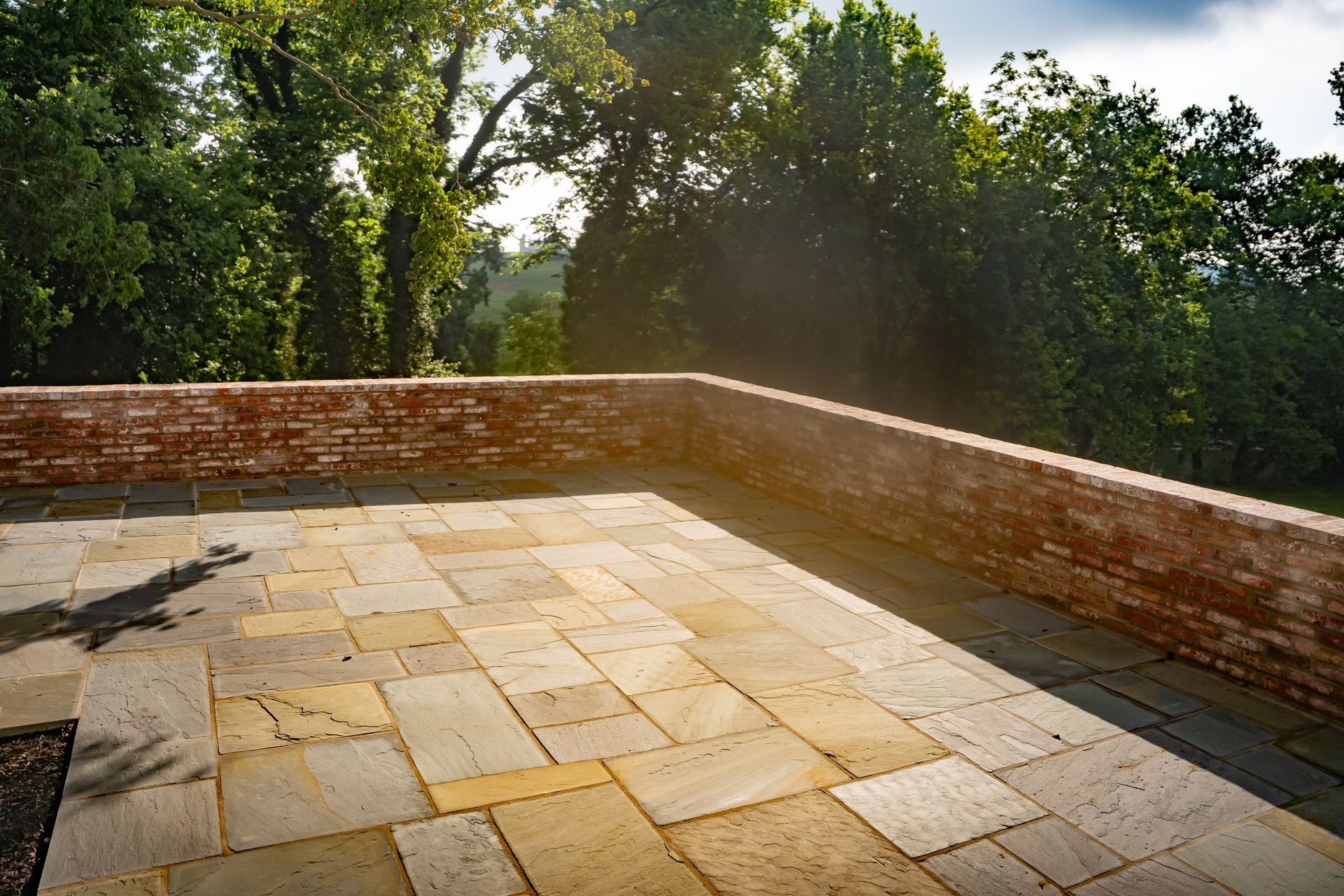 Stone patio terrace with low brick wall and trees beyond, lit by warm sunlight