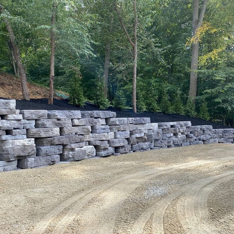 Stacked stone retaining wall along a curving dirt road in a wooded area.