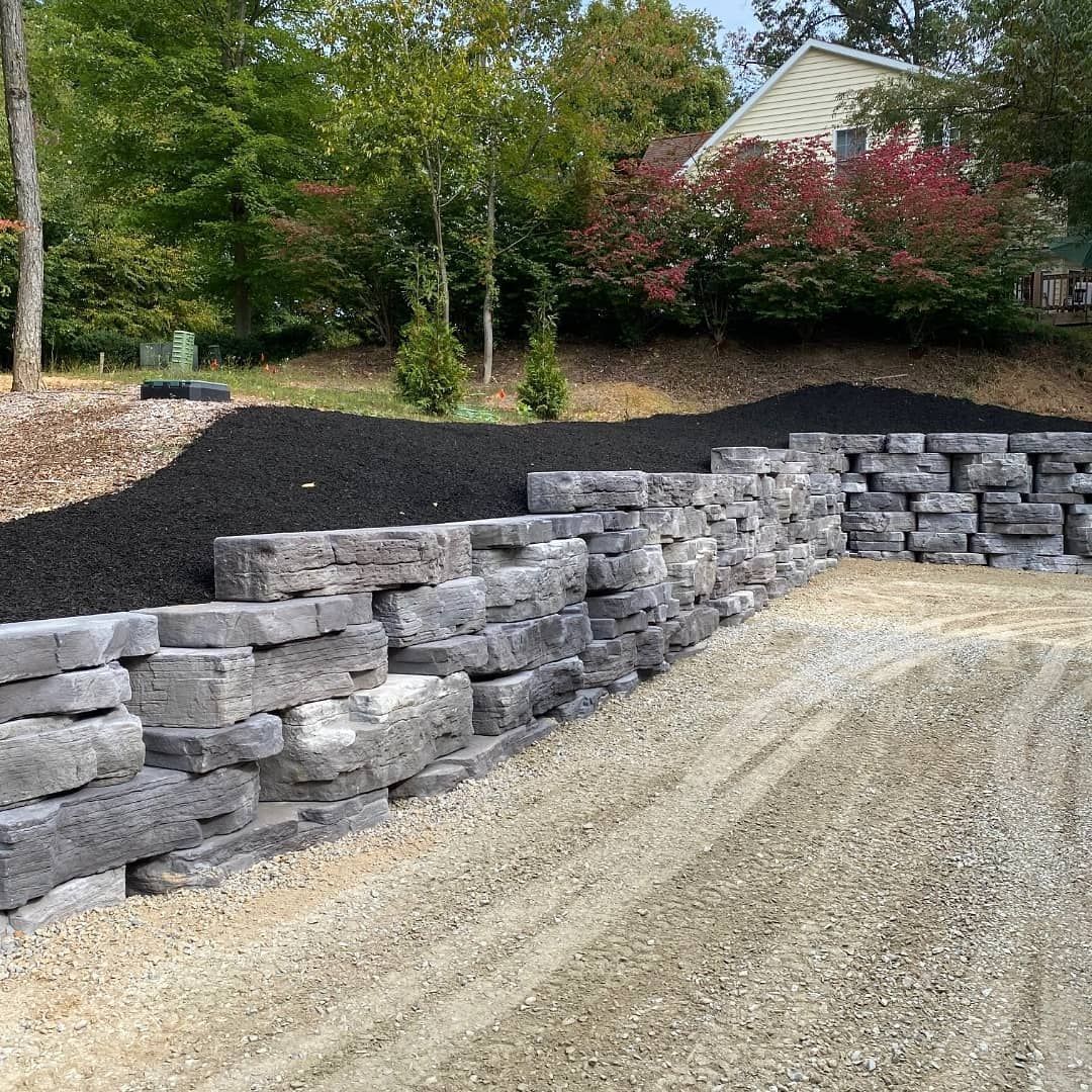 Curved gray retaining wall beside gravel driveway, with landscaped yard and trees in the background