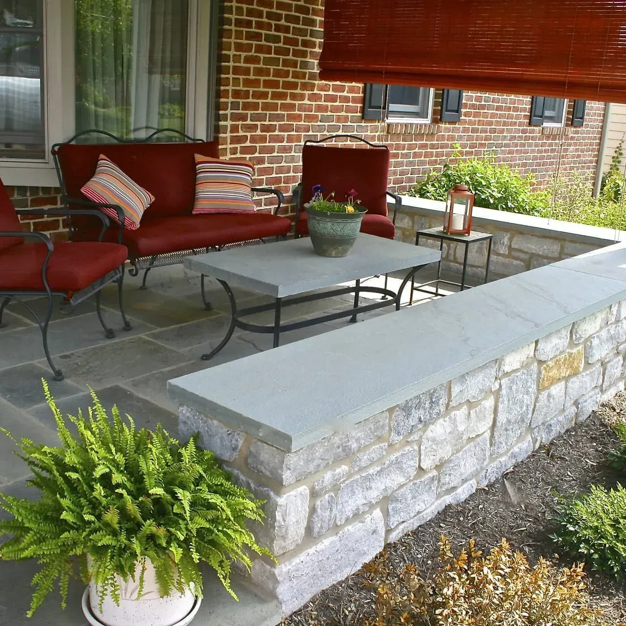 Covered stone patio with red cushioned chairs, gray table, potted plants, and brick wall backdrop