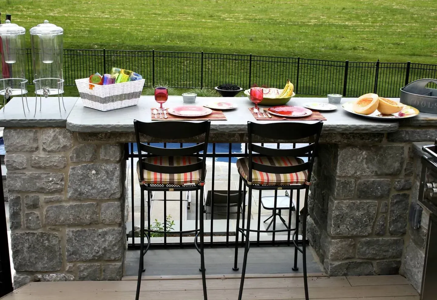 Outdoor stone patio bar with two stools, a tray of snacks, and drinks overlooking a green lawn