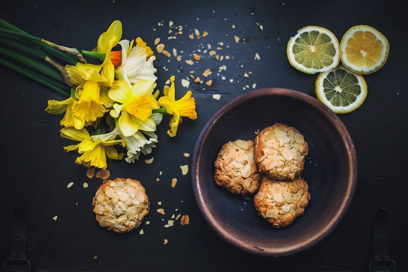 Daffodils and lime slices beside bowls of crumb-coated cookies on a dark surface
