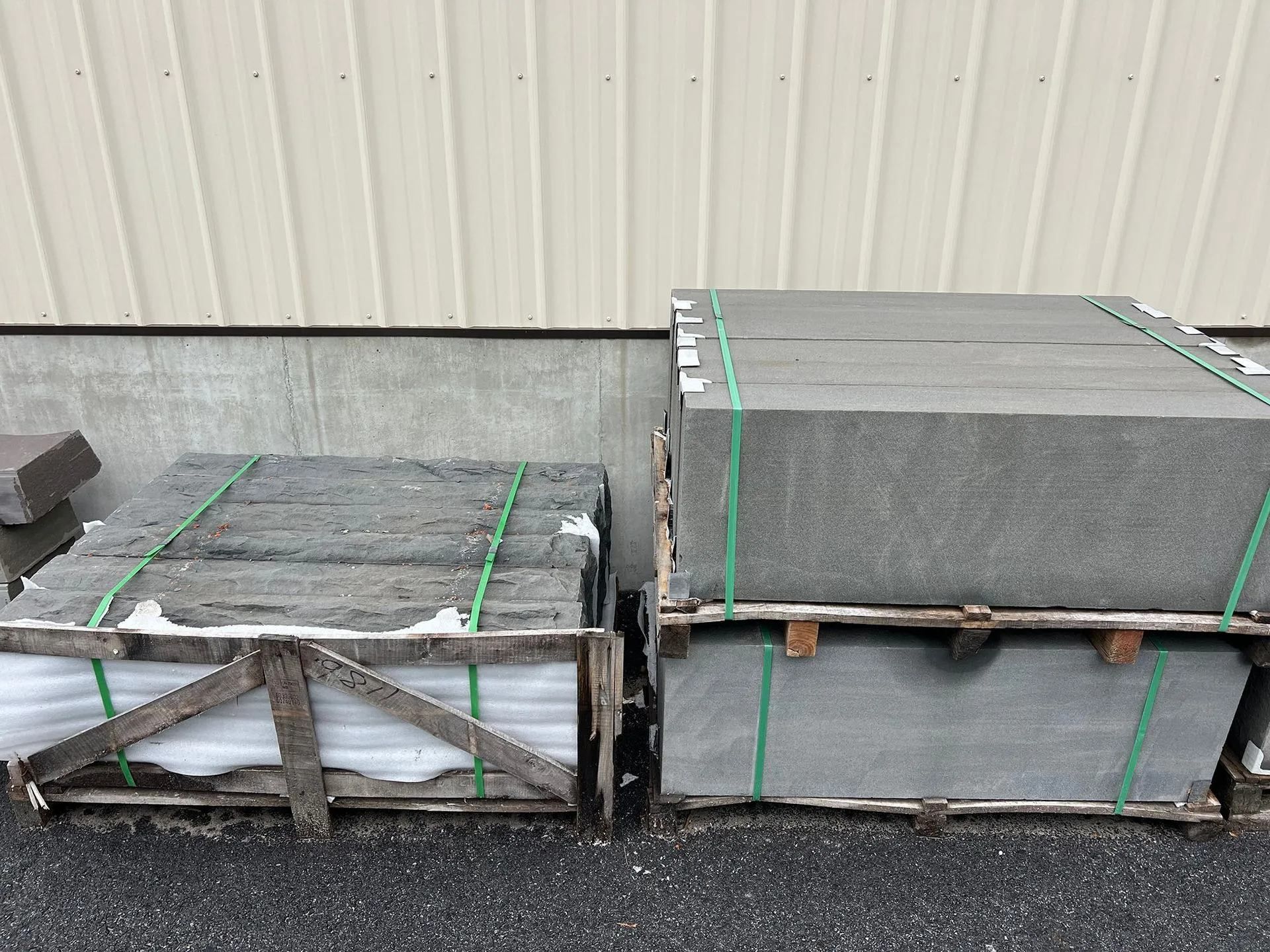 Stacked pallets of gray stone slabs strapped and wrapped in plastic against a corrugated wall.