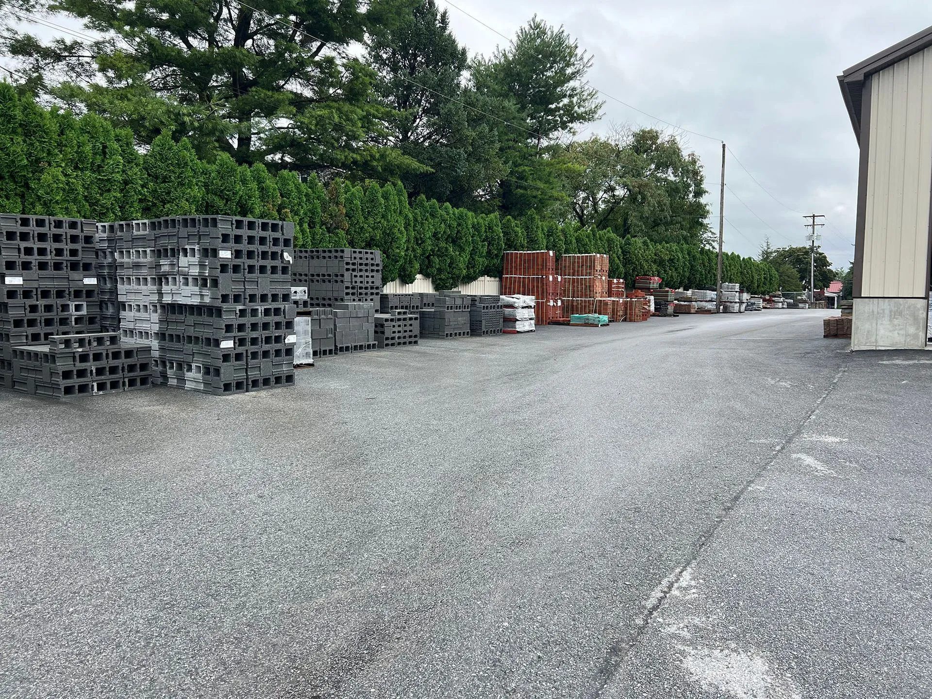 Gravel yard with stacked cinder blocks, pallets, and a warehouse building on a cloudy day