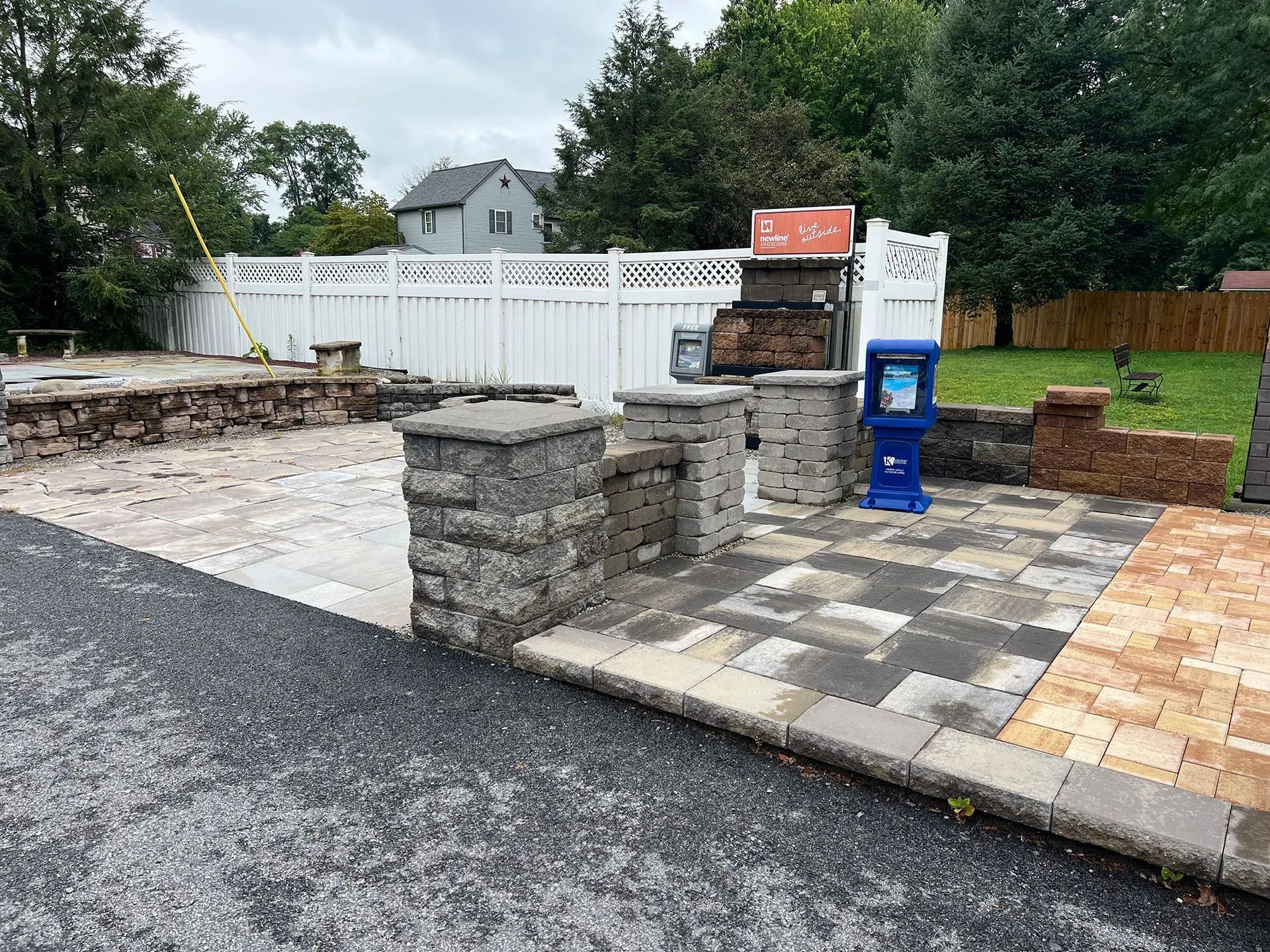 Partially paved outdoor area with stone columns, a blue mailbox, and a fenced yard behind it
