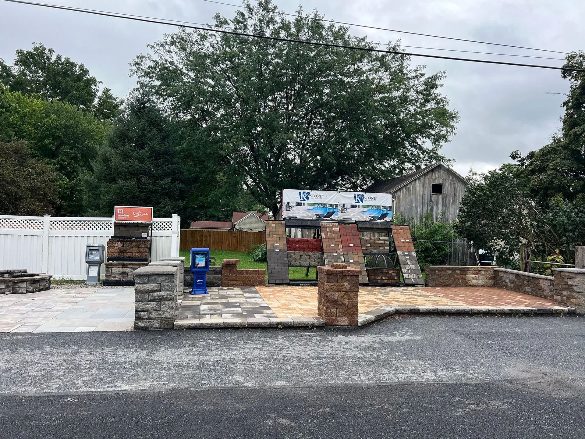 Outdoor roadside display of stacked brick samples and signs, with trees and a small shed in back.