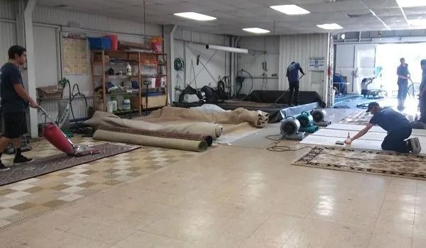 People rolling out large tan rugs in a workshop with shelves and tools in the background